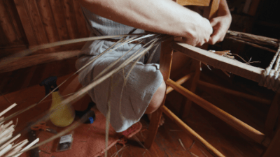 Person weaving sitting on a chair with a loom in a woodworking shop.