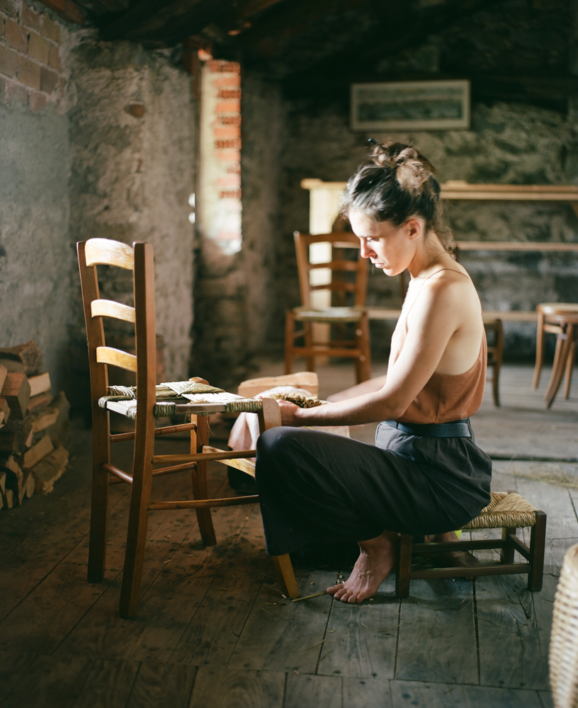 A woman sitting on a small wooden chair in an rustic room, looking down at a small object or book in her hands. The room has stone and brick walls and a wooden floor, with a window allowing natural light to illuminate her face and shoulders.