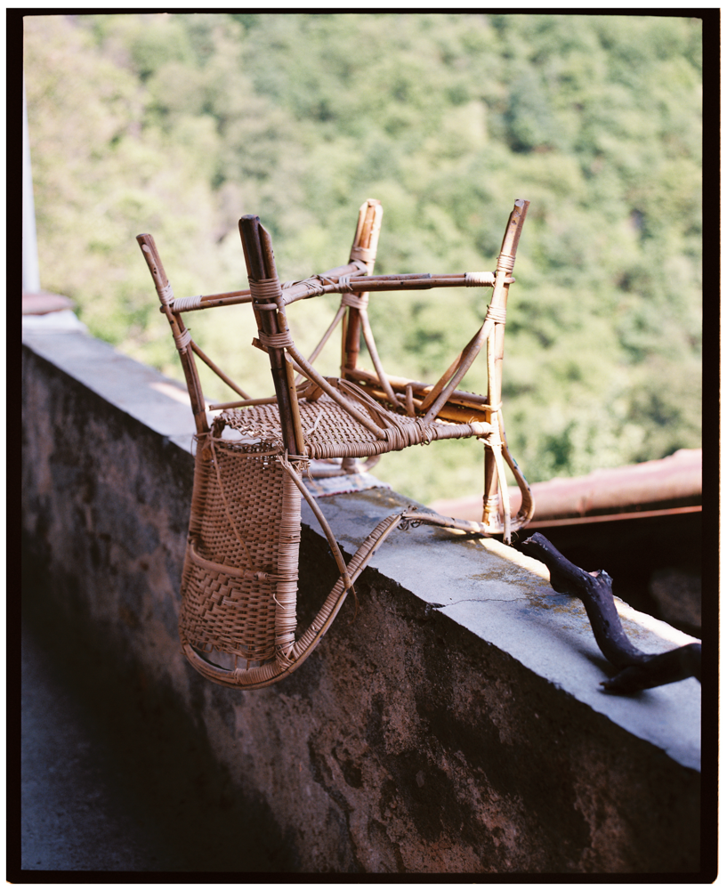 Two wicker chairs stacked upside down on a stone balcony ledge with a blurred green treeline in the background.