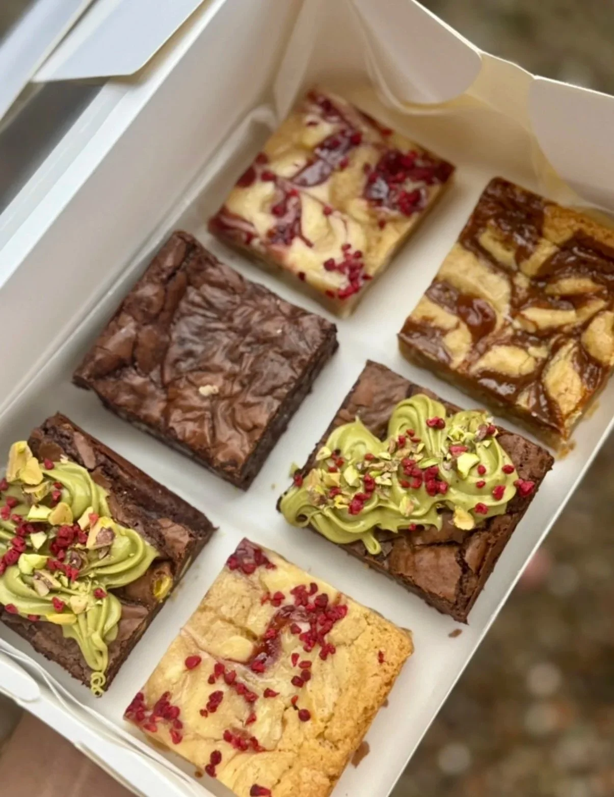 A box of six assorted brownies and blondies, featuring pistachio cream brownie, raspberry cheesecake blondie , salted caramel brownie and triple chocolate brownie.