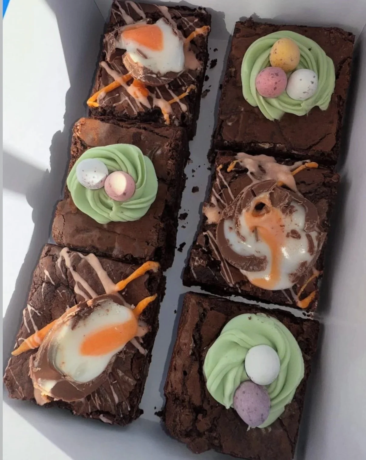 A box of six assorted brownies and blondies, featuring, cream egg brownie with fondant swirl, buttercream nest brownie with mini egg topping.