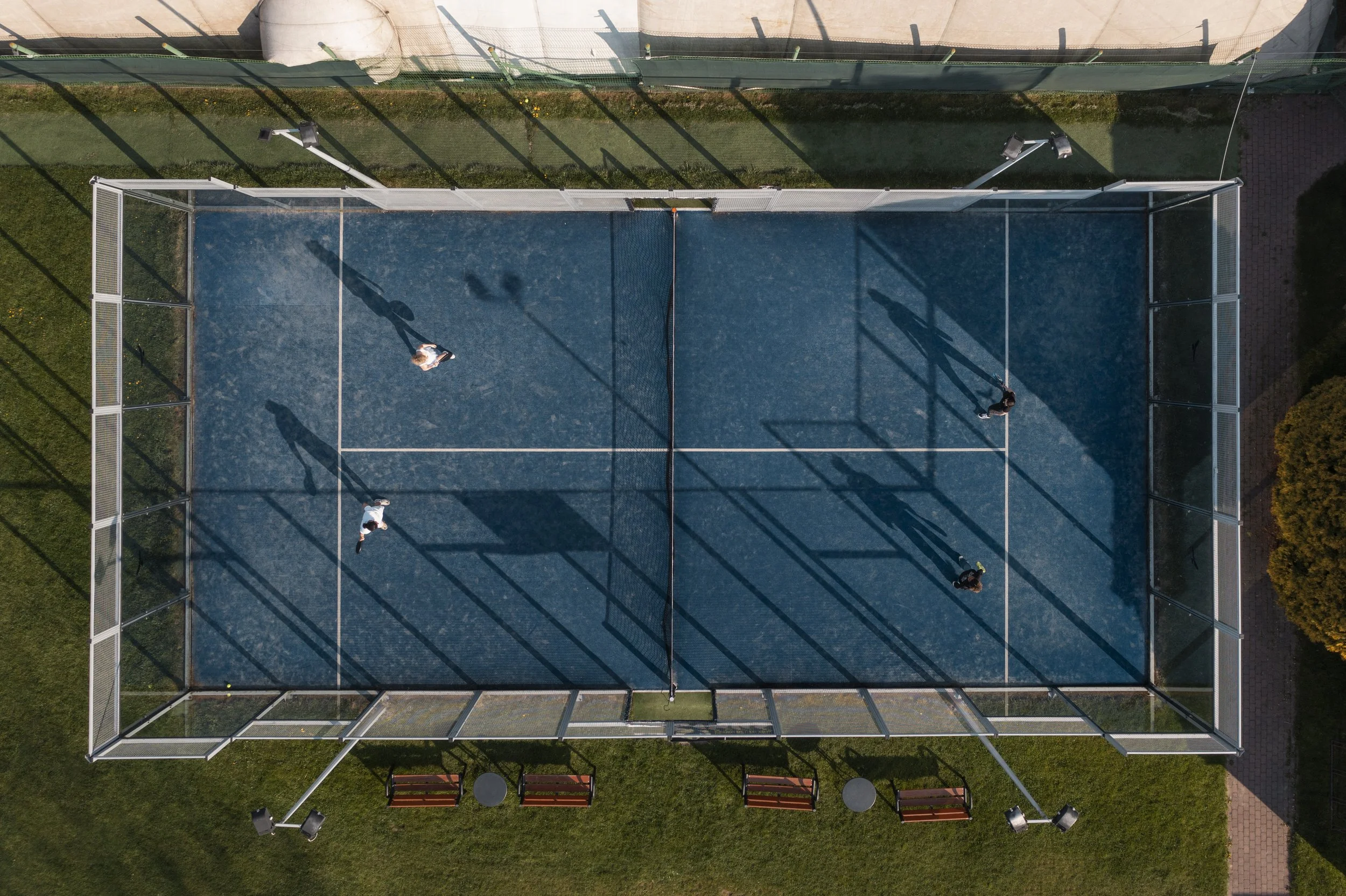 An aerial view of a blue tennis court with four people playing, surrounded by a chain-link fence, benches, and outdoor lighting.