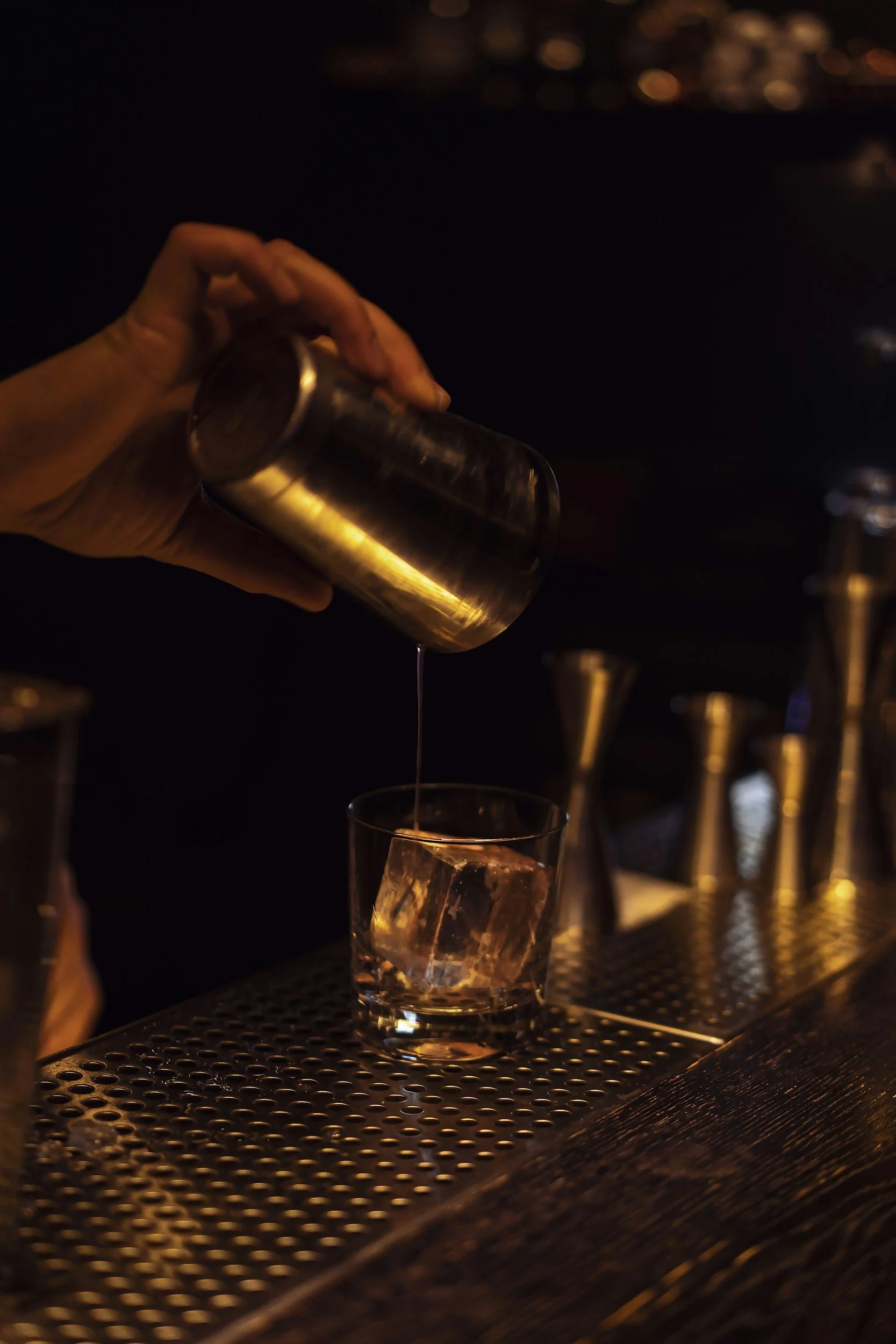 A bartender's hand is pouring a drink from a metal shaker into a tall glass filled with ice at a dimly lit bar.