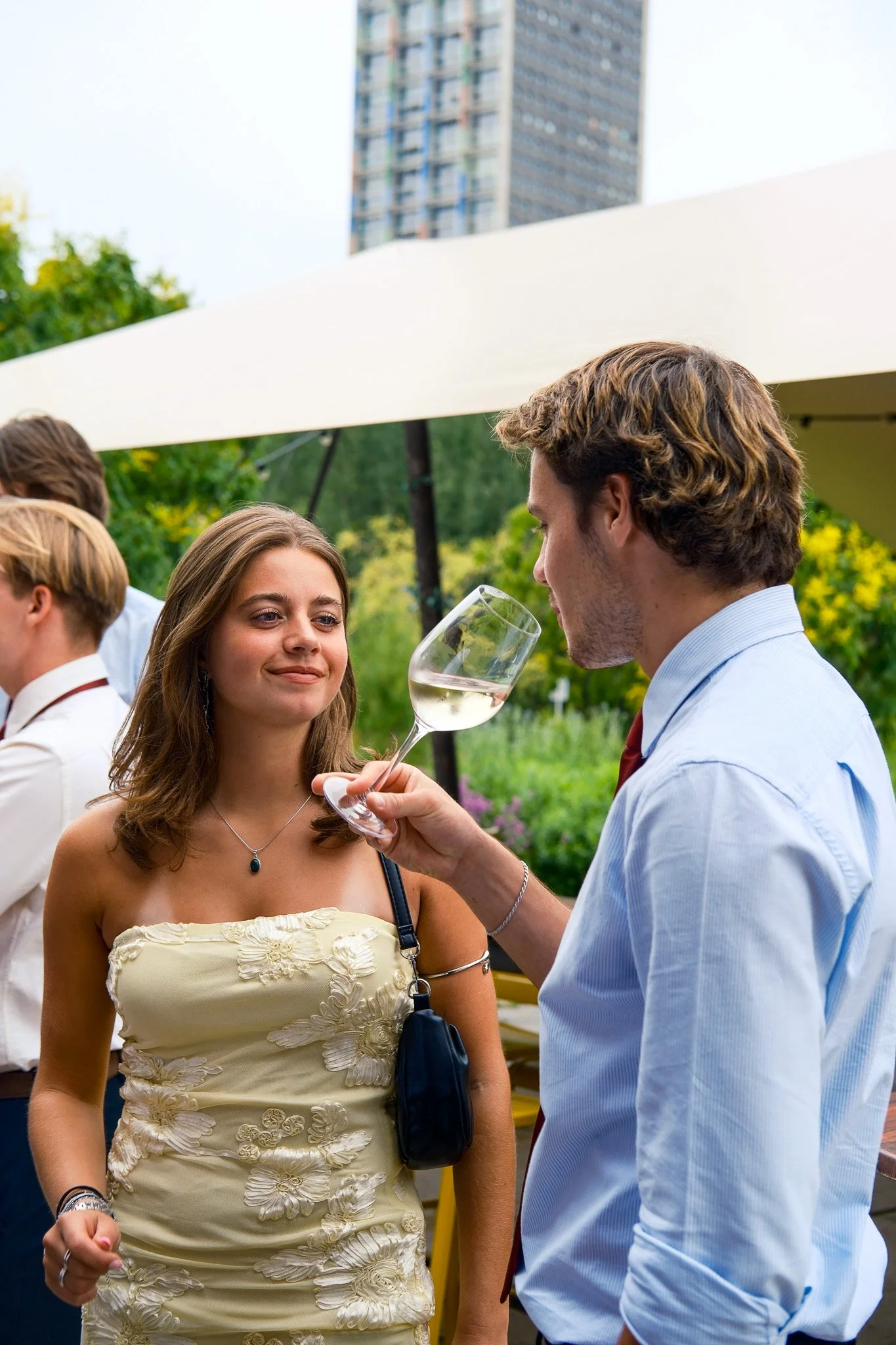 Man en vrouw op een feestje, de man schenkt wijn in het glas van de vrouw, ze glimlachen. Mensen onder een witte tent met groen landschap en een hoge gebouw op de achtergrond.