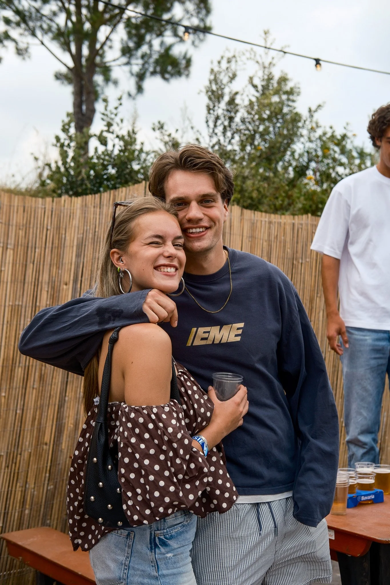 Twee mensen knuffelen en lachen op een feestje of sociale samenkomst. Meisje met een polkadot blouse en grote oorbellen, jongen met een donkerblauwe trui en gestreepte shorts.