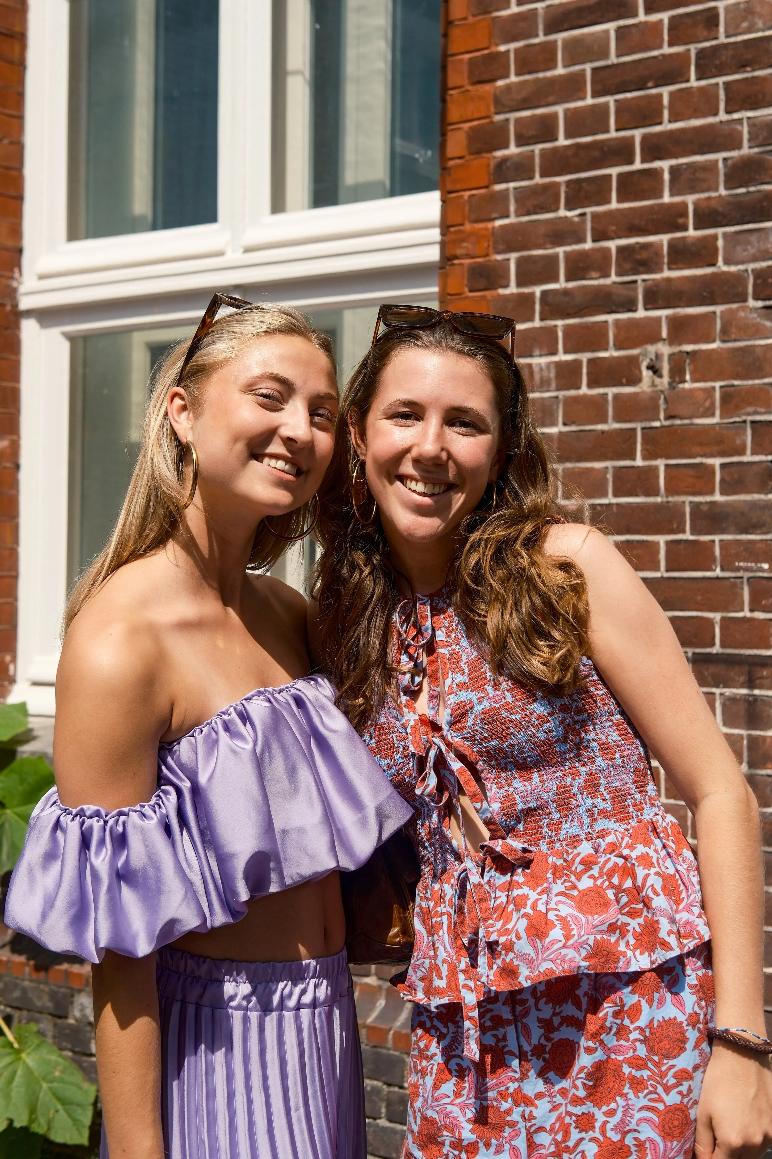 Twee jonge vrouwen staan dicht bij elkaar buiten, lachend, voor een bakstenen muur en een raam, in de zon.