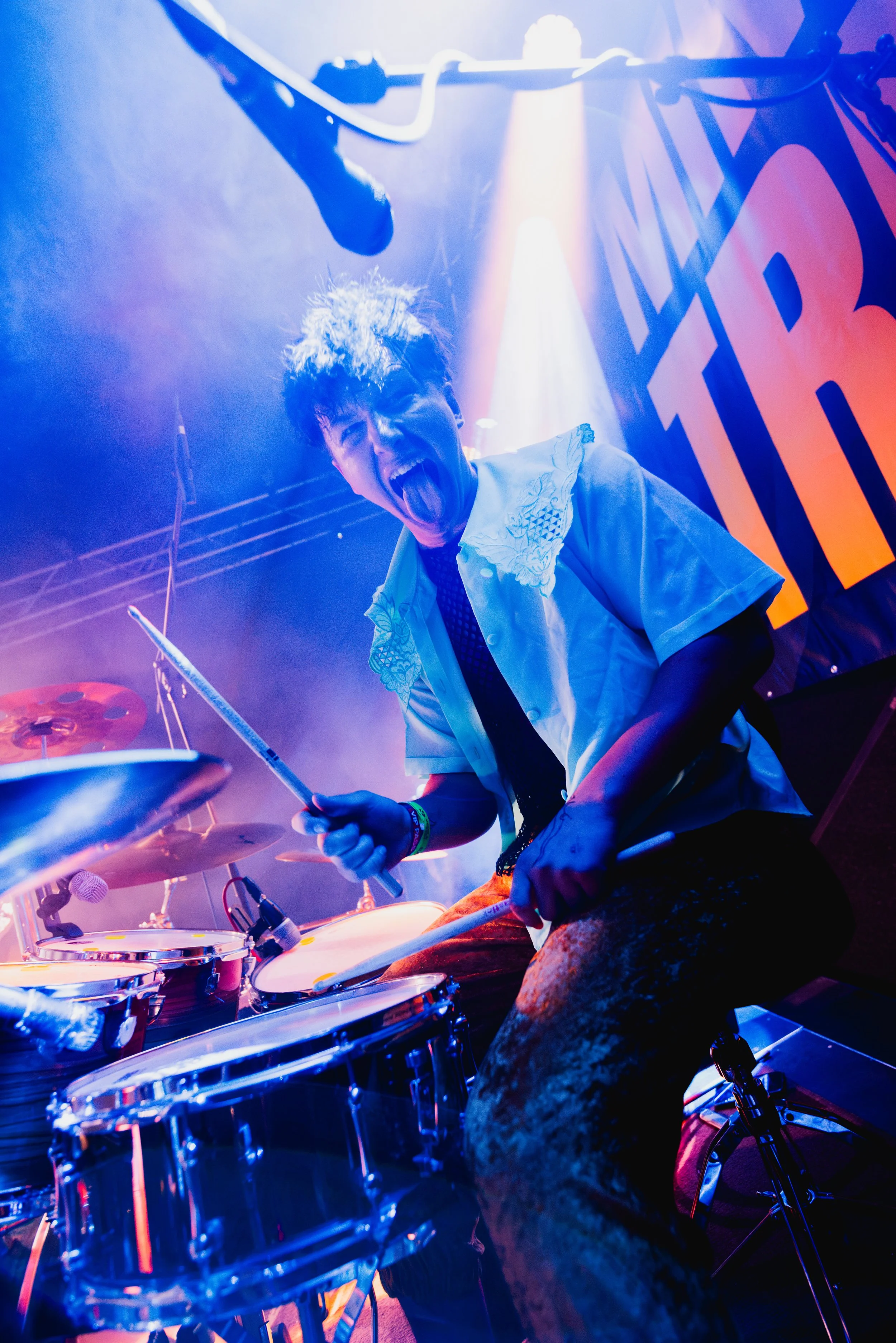 A drummer playing drums under colorful stage lighting, sticking tongue out, wearing a white embroidered shirt, with a large sign or banner with bold letters in the background.