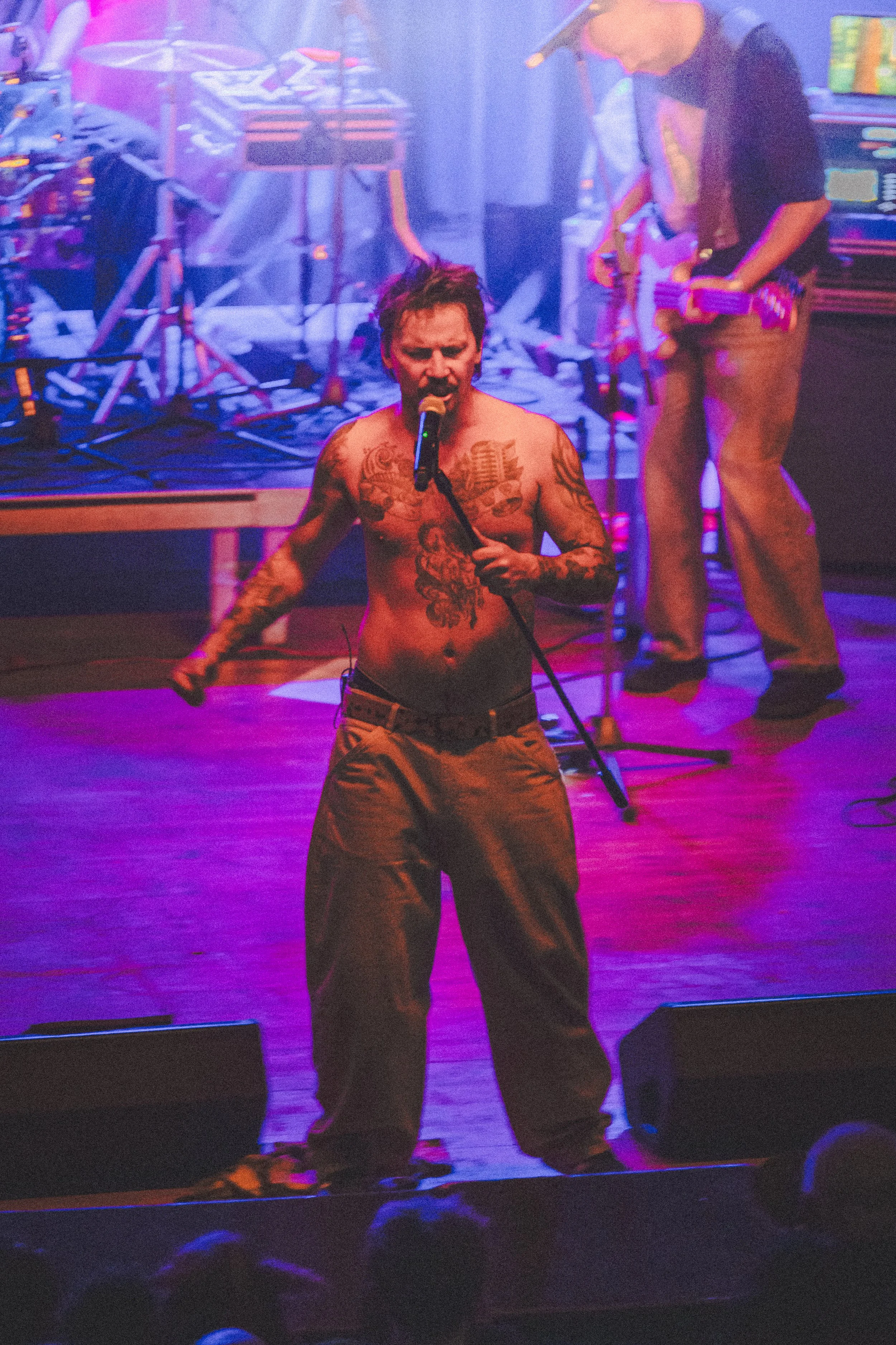 A shirtless male performer with tattoos singing into a microphone on stage with purple lighting, accompanied by a person in the background holding a guitar.