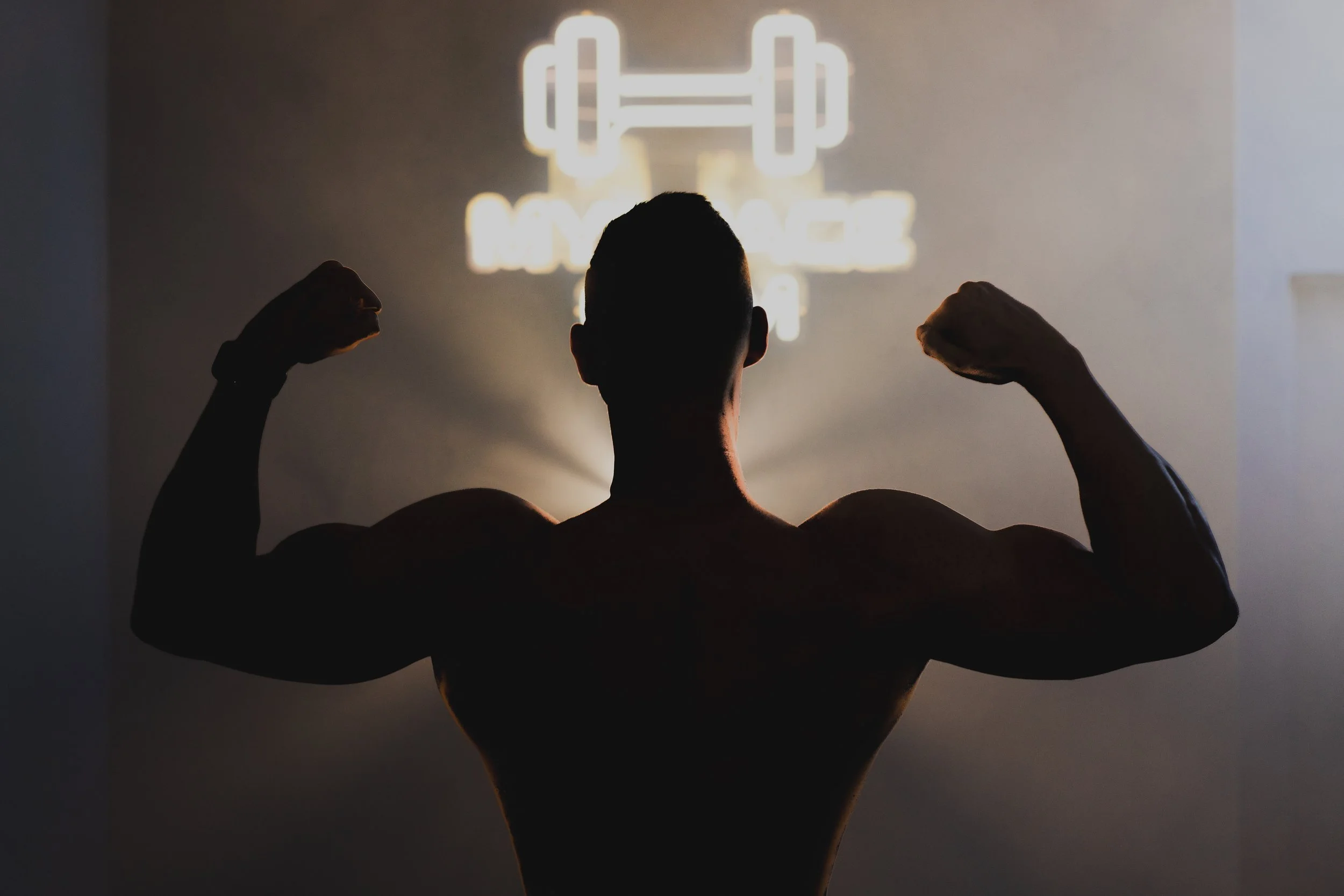A person with a muscular physique flexing their biceps in front of a wall with a neon motivational fitness quote that is blurry and difficult to read.