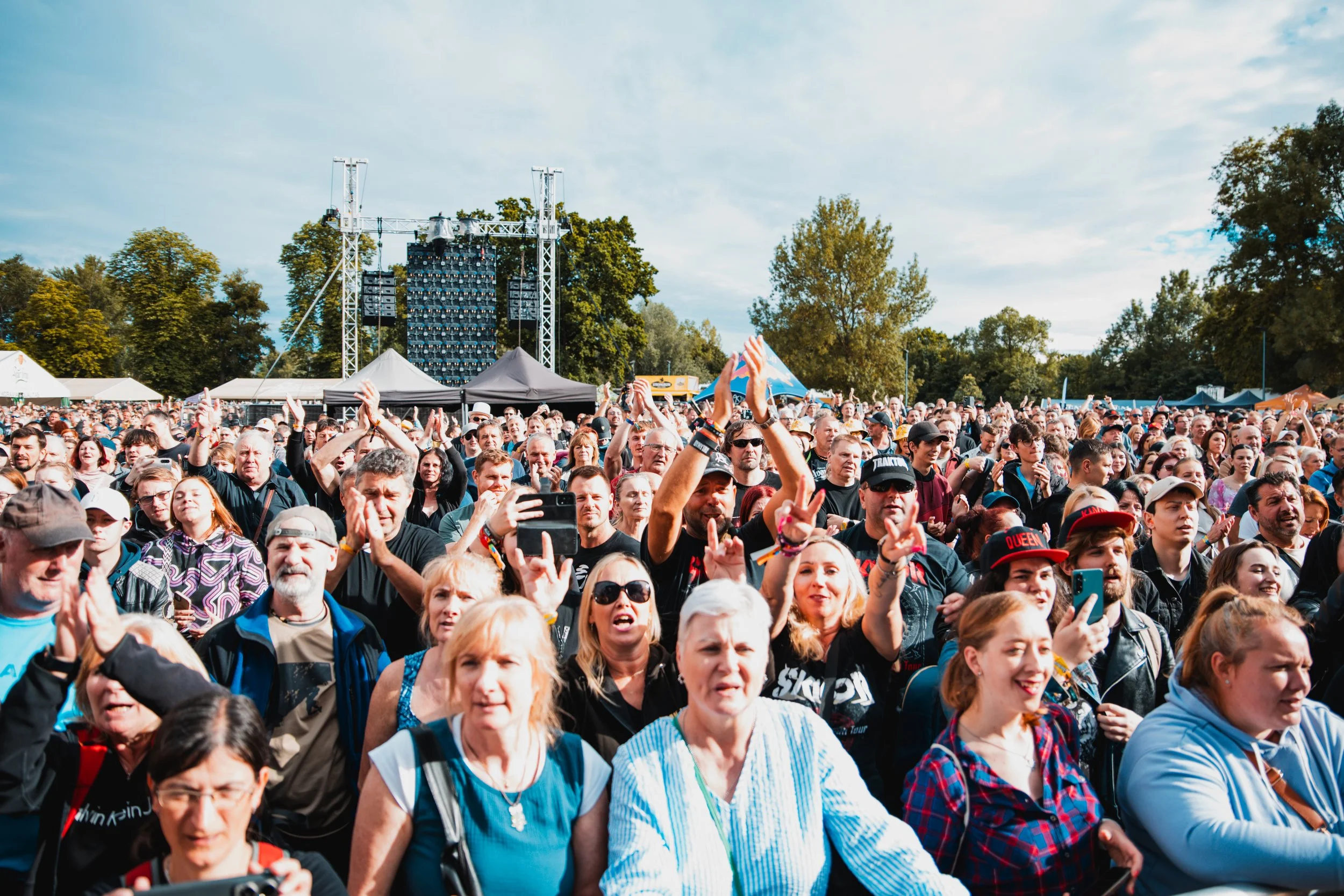 Crowd of people at an outdoor concert or festival, with a stage and large speakers in the background, surrounded by trees and tents.