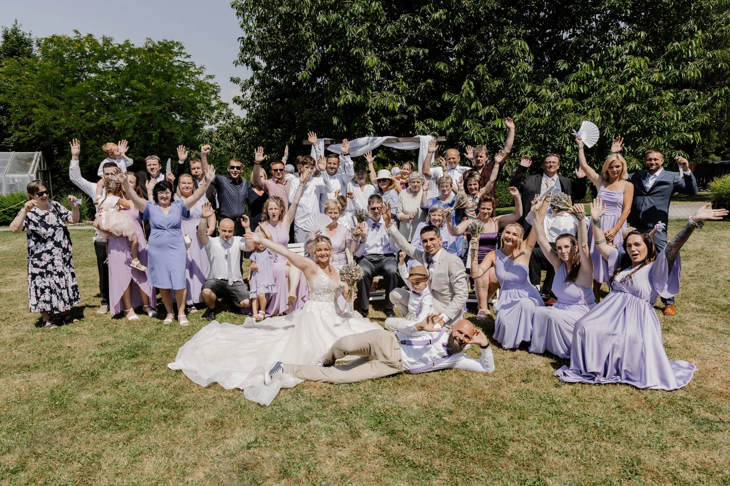 A large group of people, including men, women, and children, gathered outdoors on a sunny day for a celebration, with many raising their hands and smiling, in front of green trees and a clear sky.