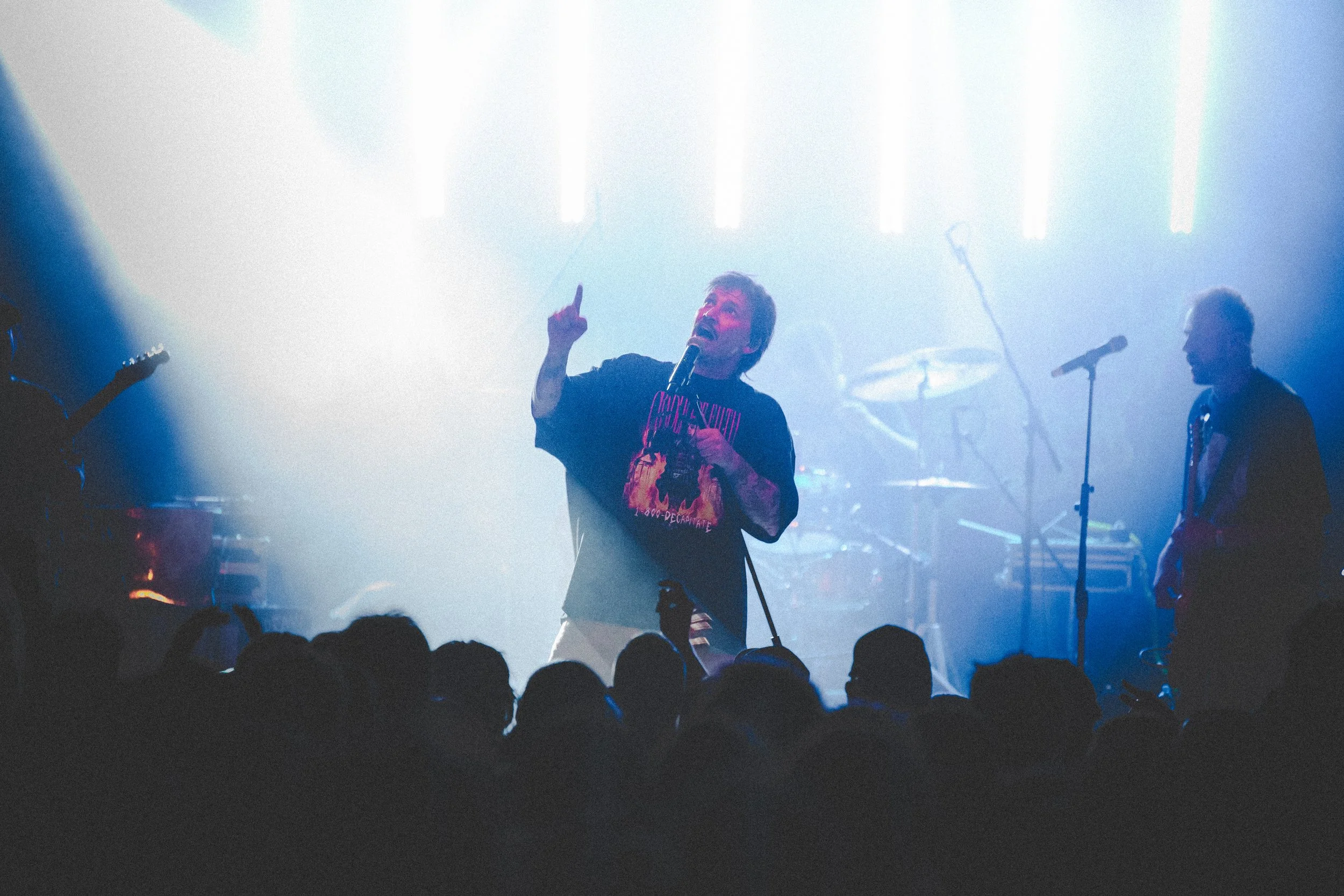 A male singer performing on stage with a microphone, wearing a dark t-shirt with pink and yellow graphics, and gesturing with one finger up. There are band members and musical instruments in the background, with bright stage lights creating a vibrant