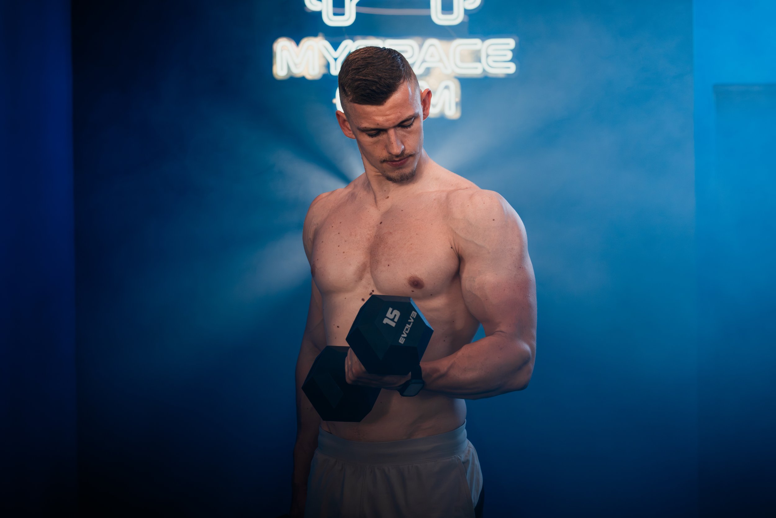 A shirtless man with short dark hair and a goatee is holding a black dumbbell with the number 15 on it in his right hand. He is standing in front of a dark blue wall with a neon sign that says 'MY SPACE MOM' in the background.