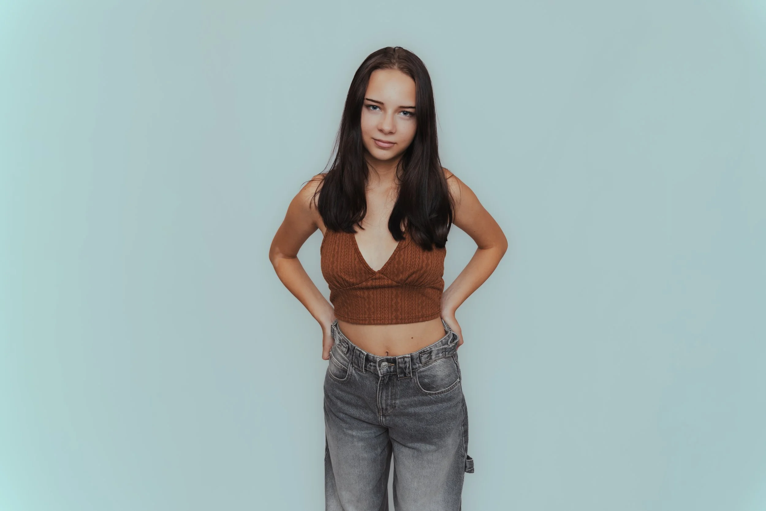 Young woman with long dark hair wearing a brown sleeveless top and gray jeans, standing with her hands on her hips against a light blue background.