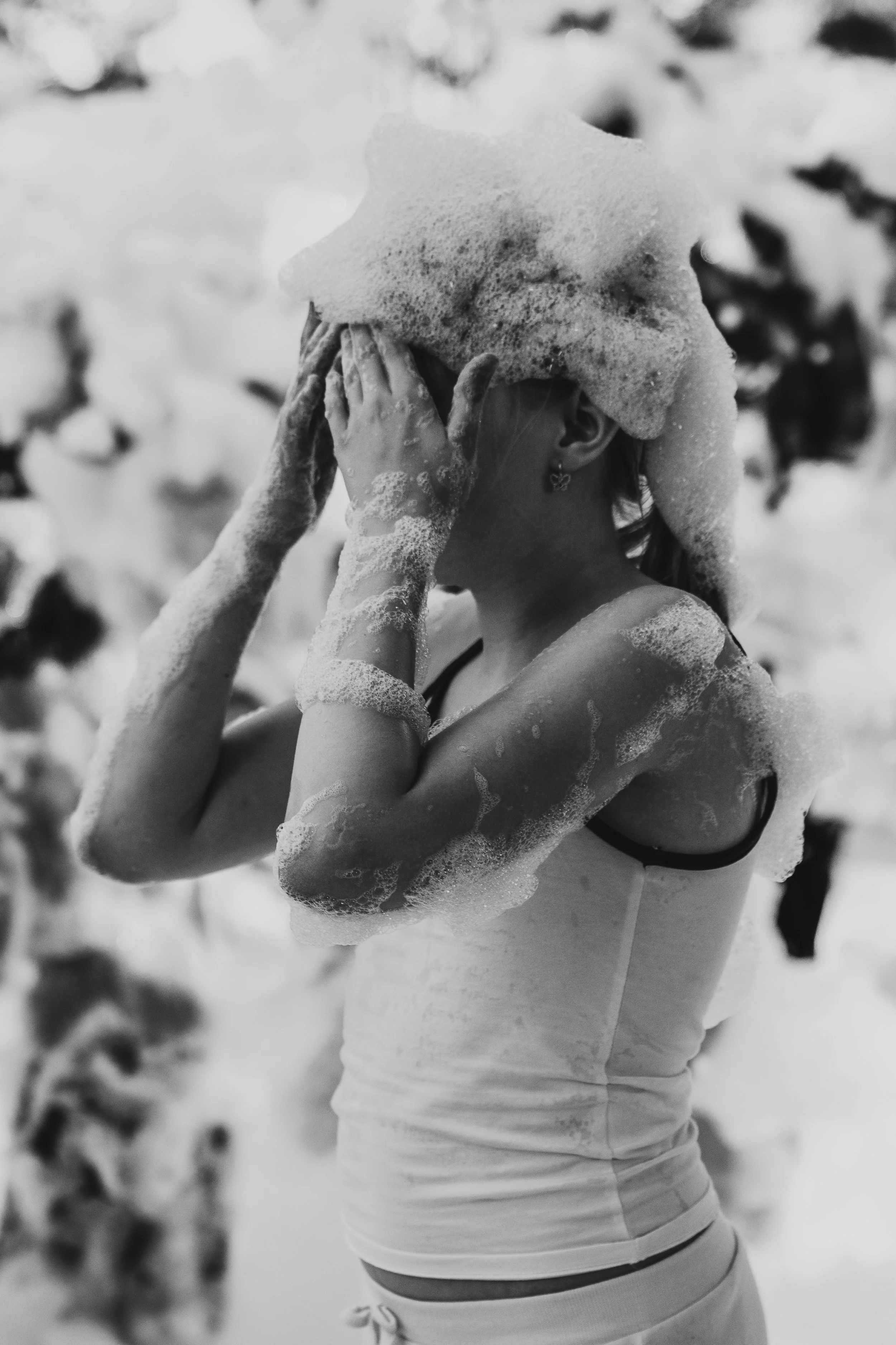 A woman covered in soap bubbles, washing her hair in a shower or bathtub, seen from the side. She has earrings and wears a tank top or swimsuit.