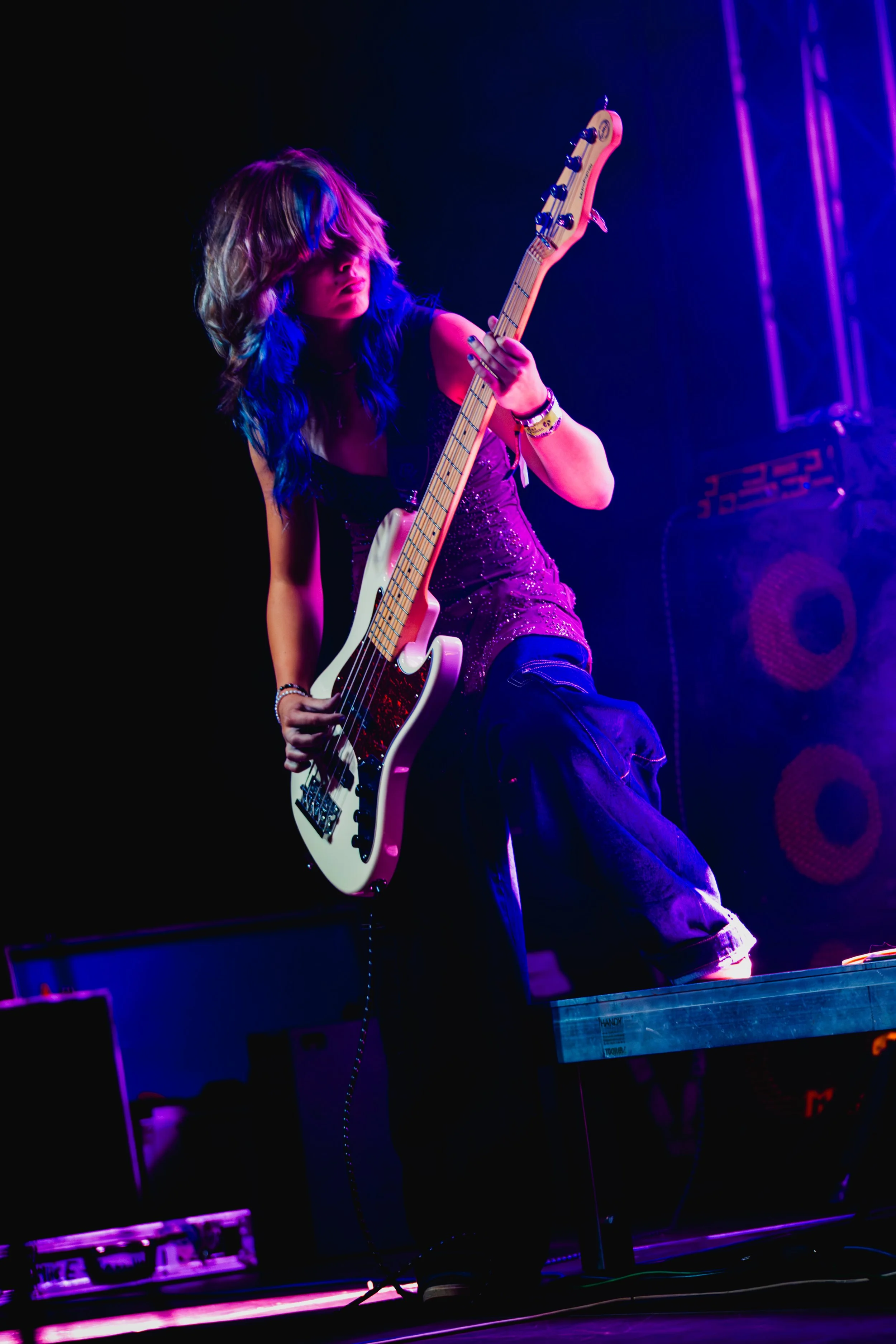 A woman with long wavy hair is playing an electric bass guitar on stage, illuminated by purple and blue stage lights, with amplifiers and keyboard in the background.
