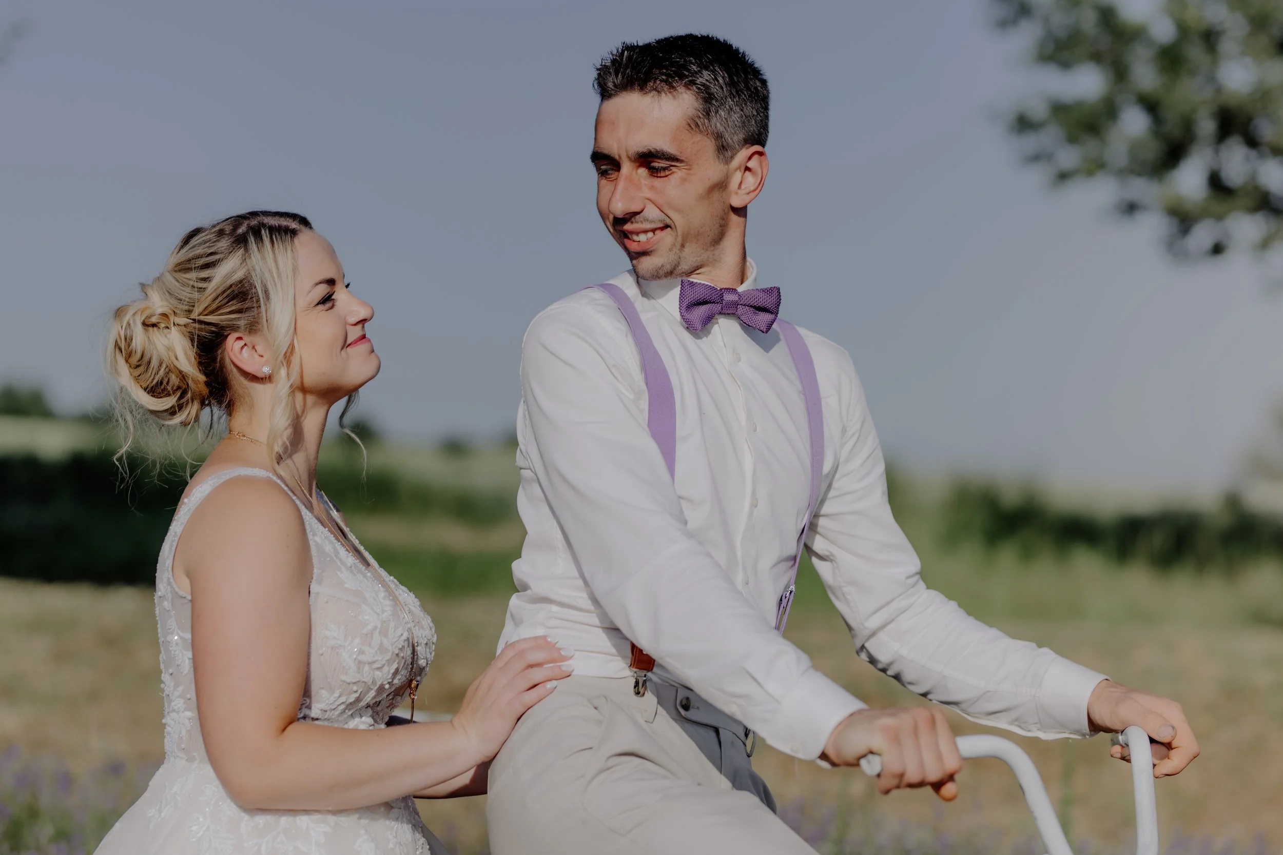 A couple riding a bicycle outdoors on a sunny day, with the woman smiling at the man and the man looking at her, against a background of green fields and a blue sky with some clouds.