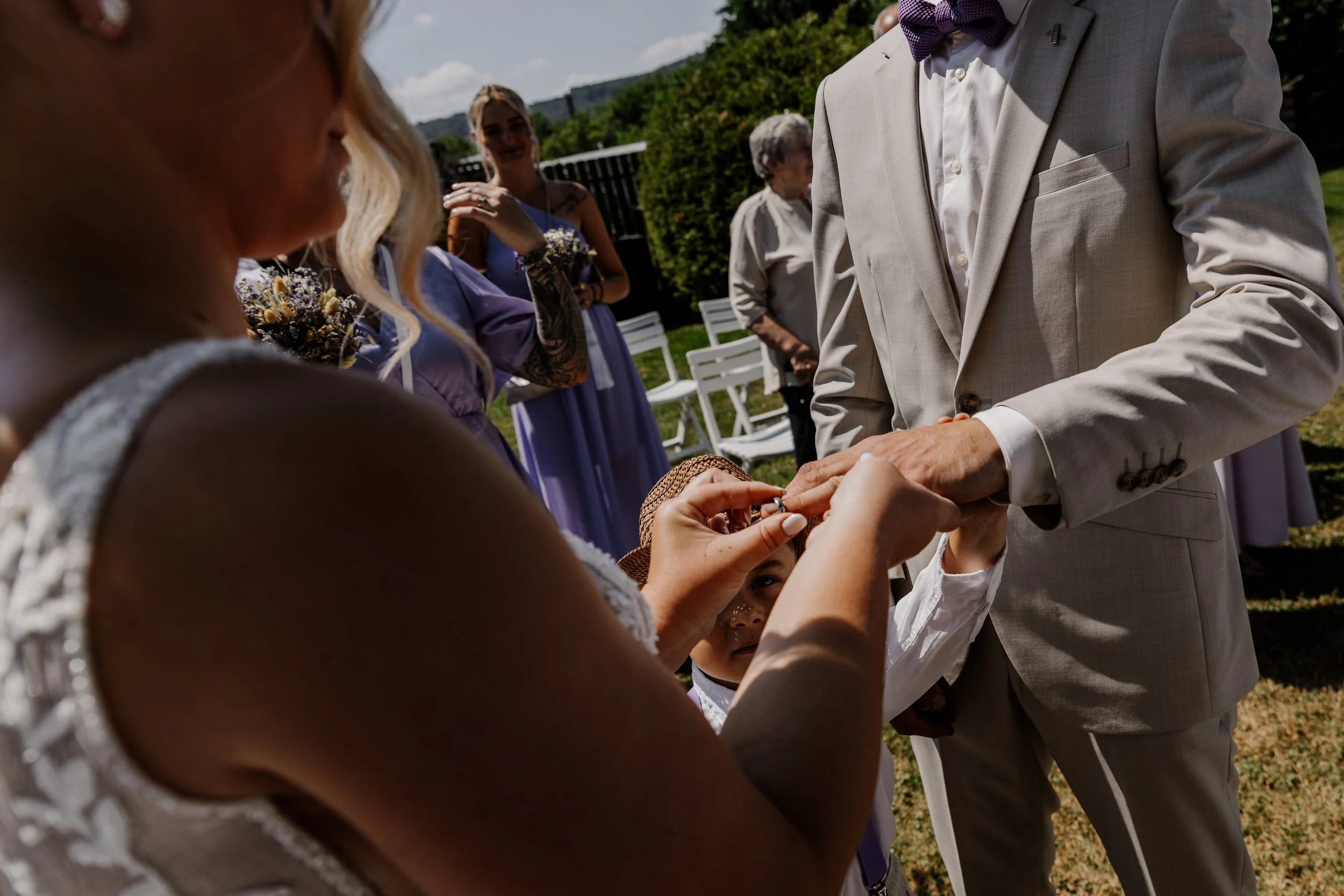 A wedding ceremony outdoors with a bride and groom exchanging rings, surrounded by bridesmaids and guests on a sunny day.