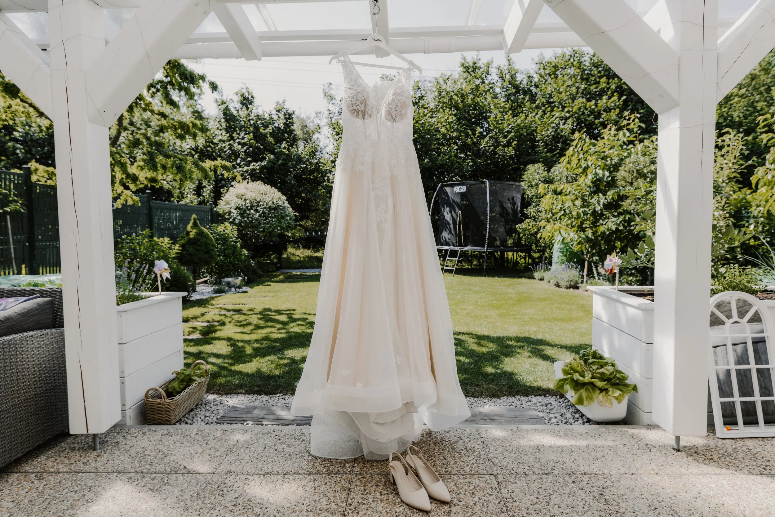 A white wedding dress hanging from a clothes hanger under a white porch, with white heels placed underneath. The scene is set outdoors in a garden area with green trees and bushes.