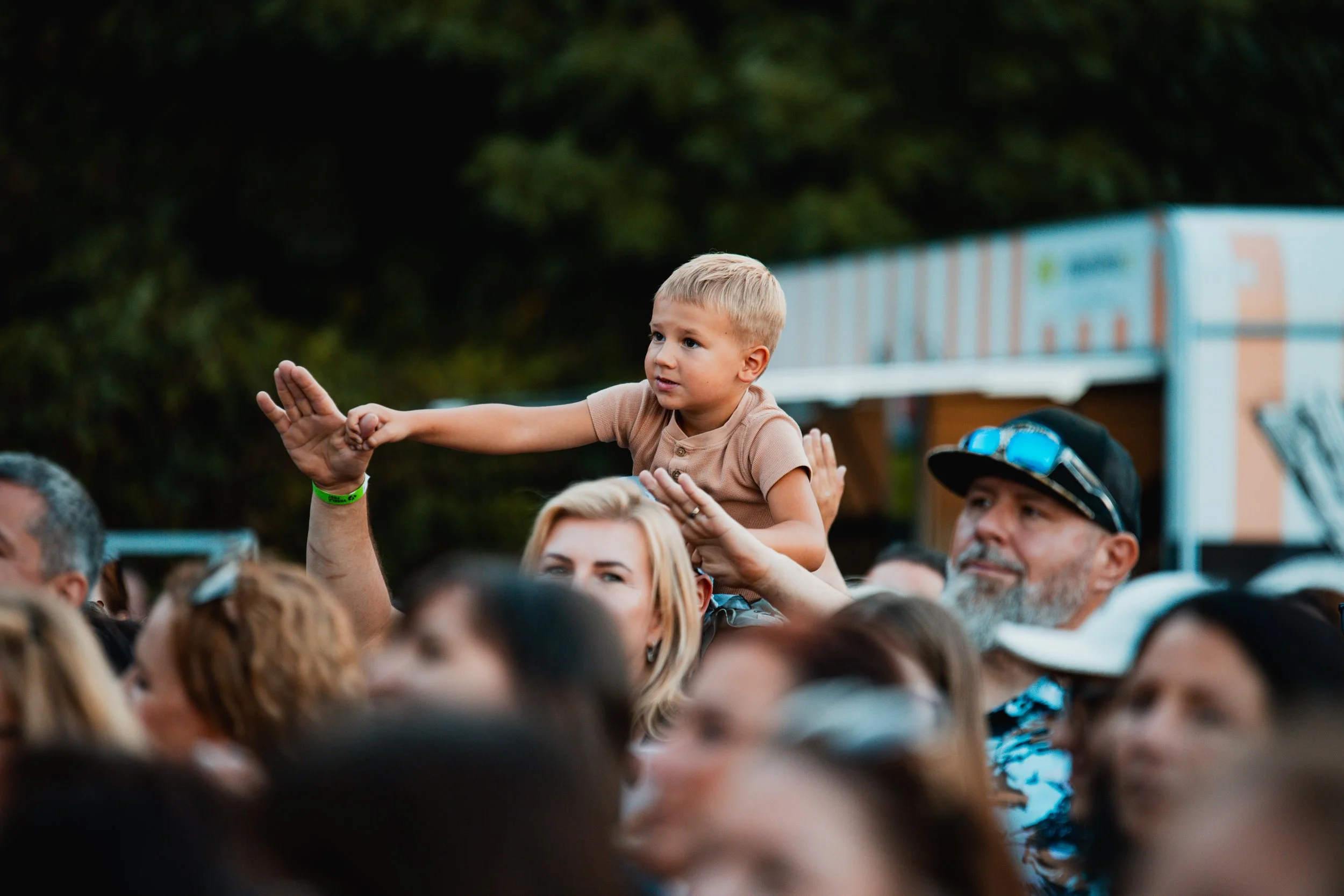 A young boy with blond hair sitting on an adult's shoulders in a crowd at an outdoor event, with people around and trees in the background.