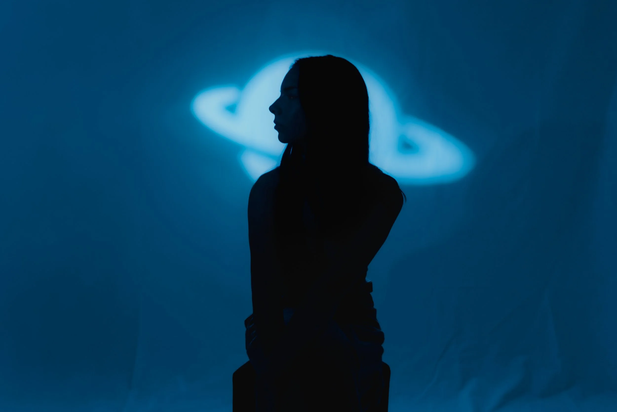 Silhouette of a woman with long hair standing against a blue wall with a glowing heart-shaped neon light in the background.