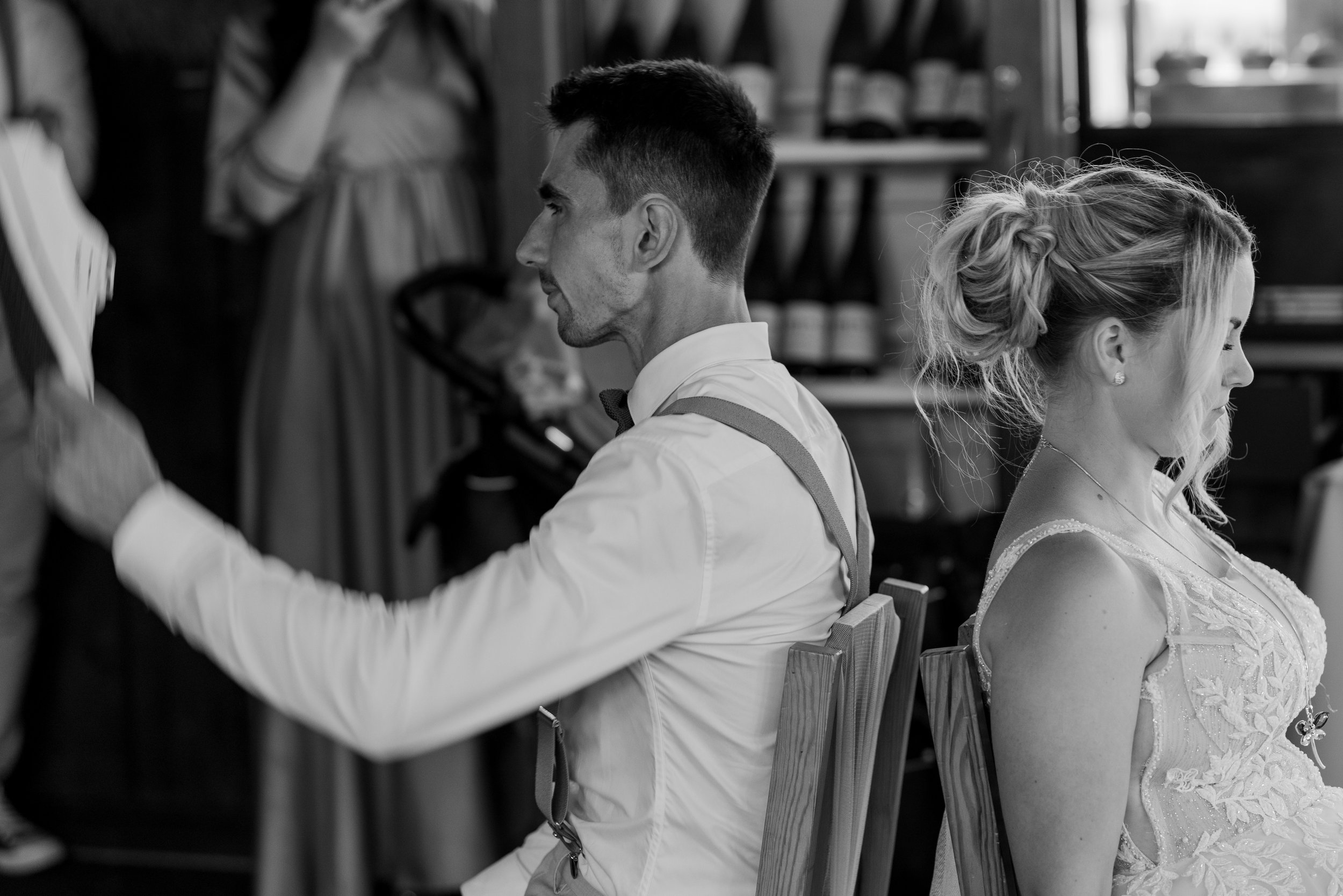 Black and white photo of a groom and bride sitting back to back, eyes closed, in a wedding venue with friends and family in the background.