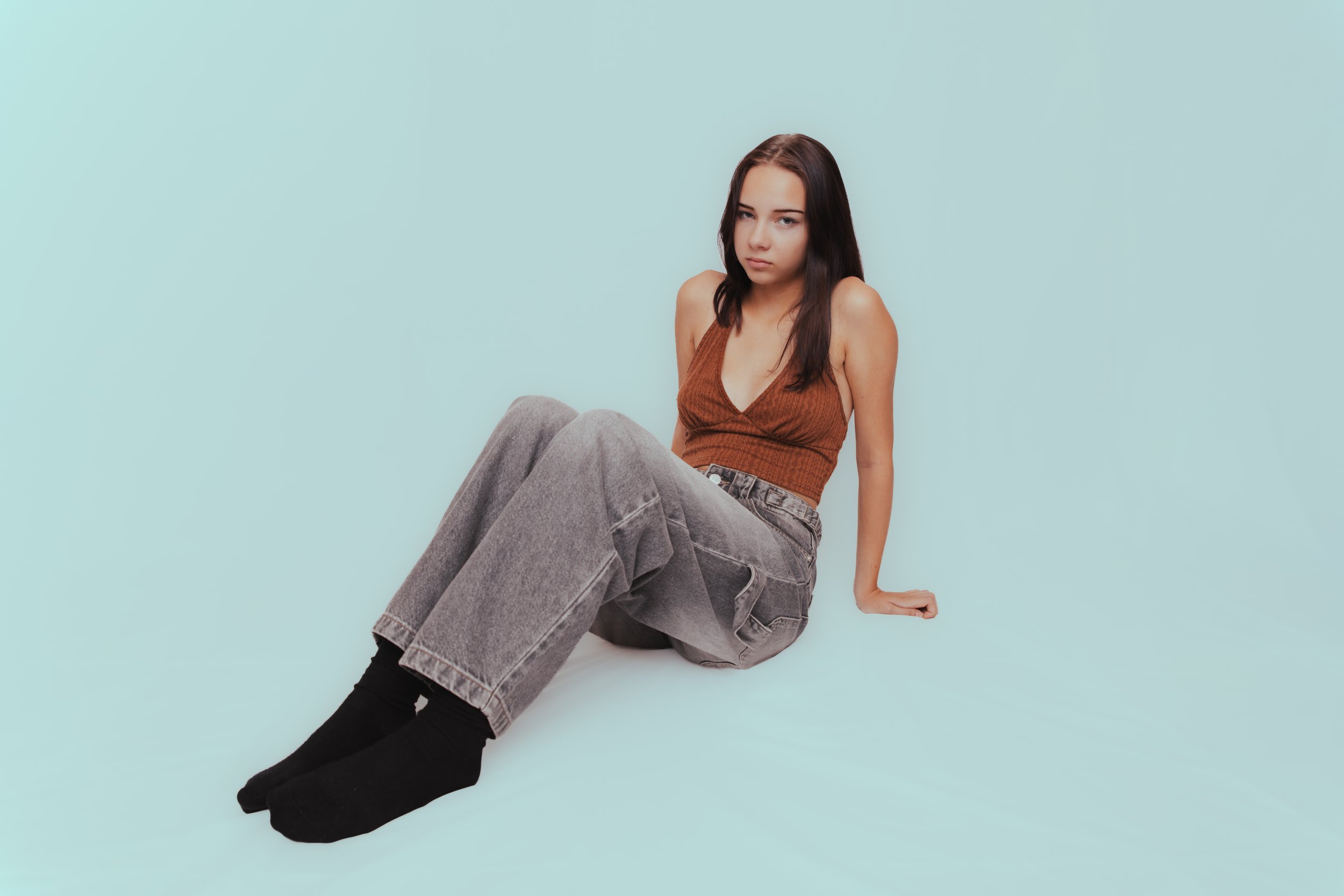 Young woman with long dark hair wearing a brown sleeveless top, gray jeans, and black socks sitting against a plain light blue background.