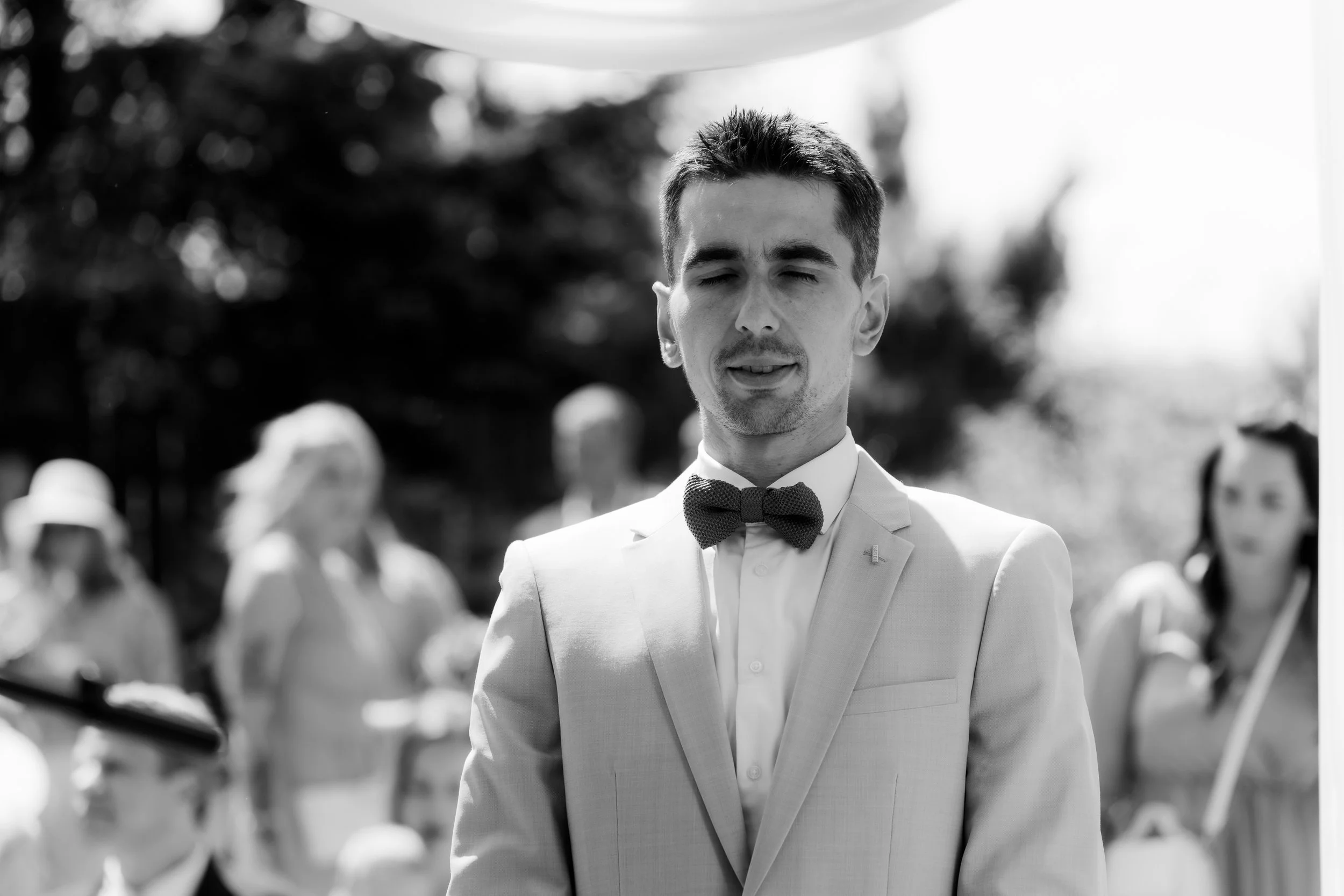 A black and white photo of a young man in formal attire, wearing a tuxedo with a bow tie, standing outdoors with a blurred background of people and trees. He has short dark hair and appears to be at a formal event.
