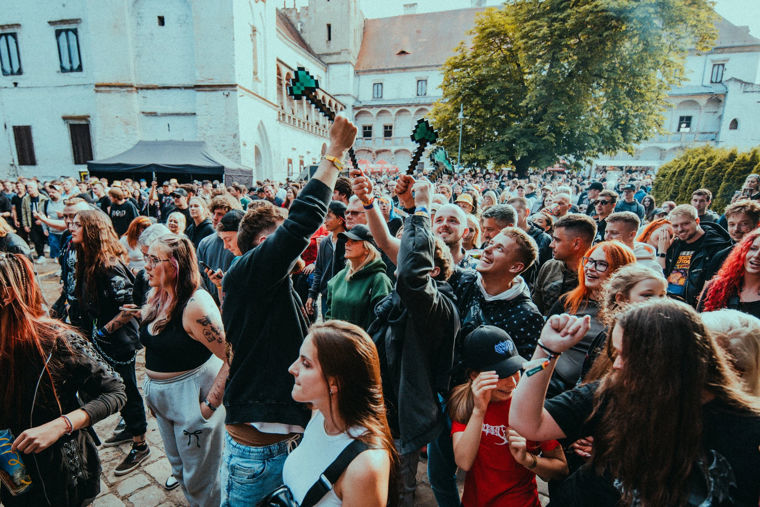Crowd of people gathered outdoors near historic buildings, some holding pixelated stone swords props, smiling and enjoying the event on a sunny day.