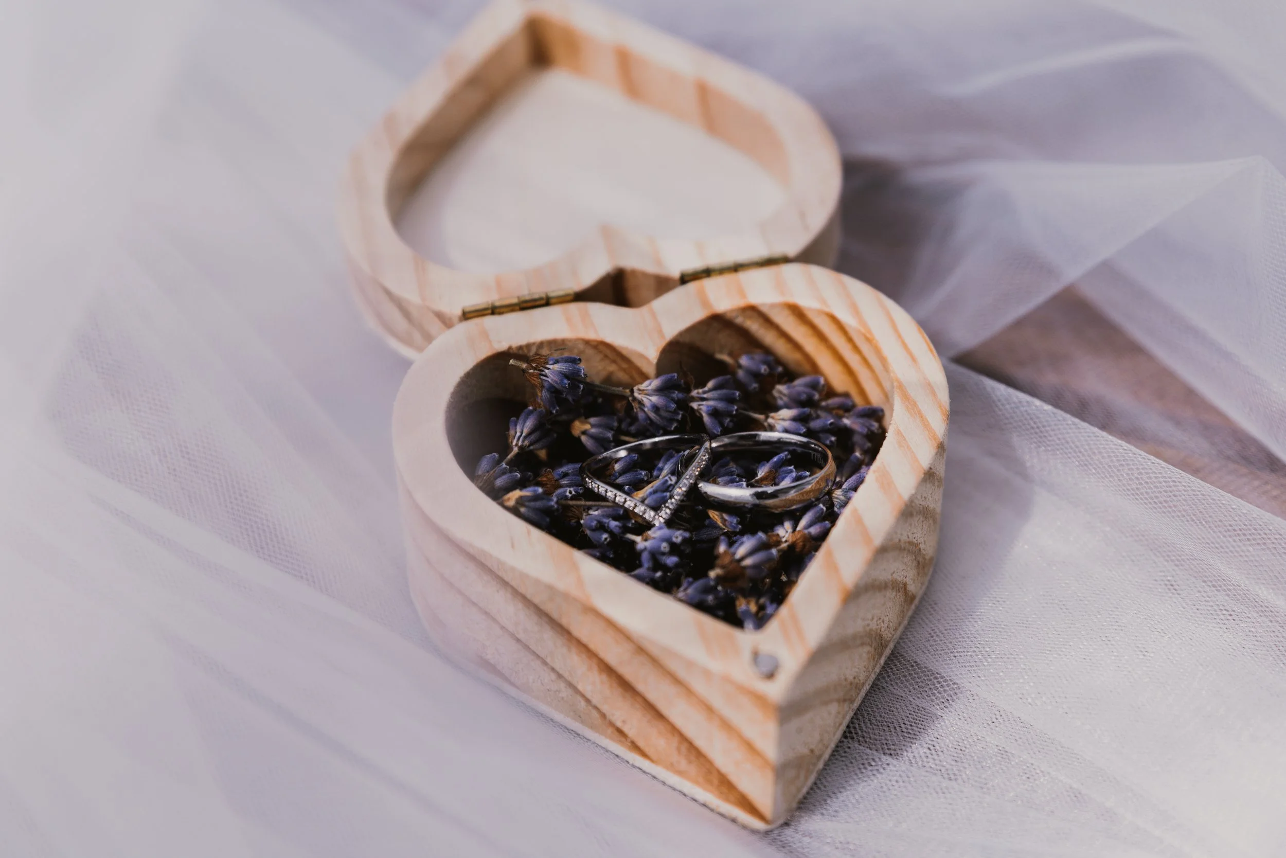 A wooden heart-shaped box filled with dried lavender flowers and small rings, placed on a white fabric surface.