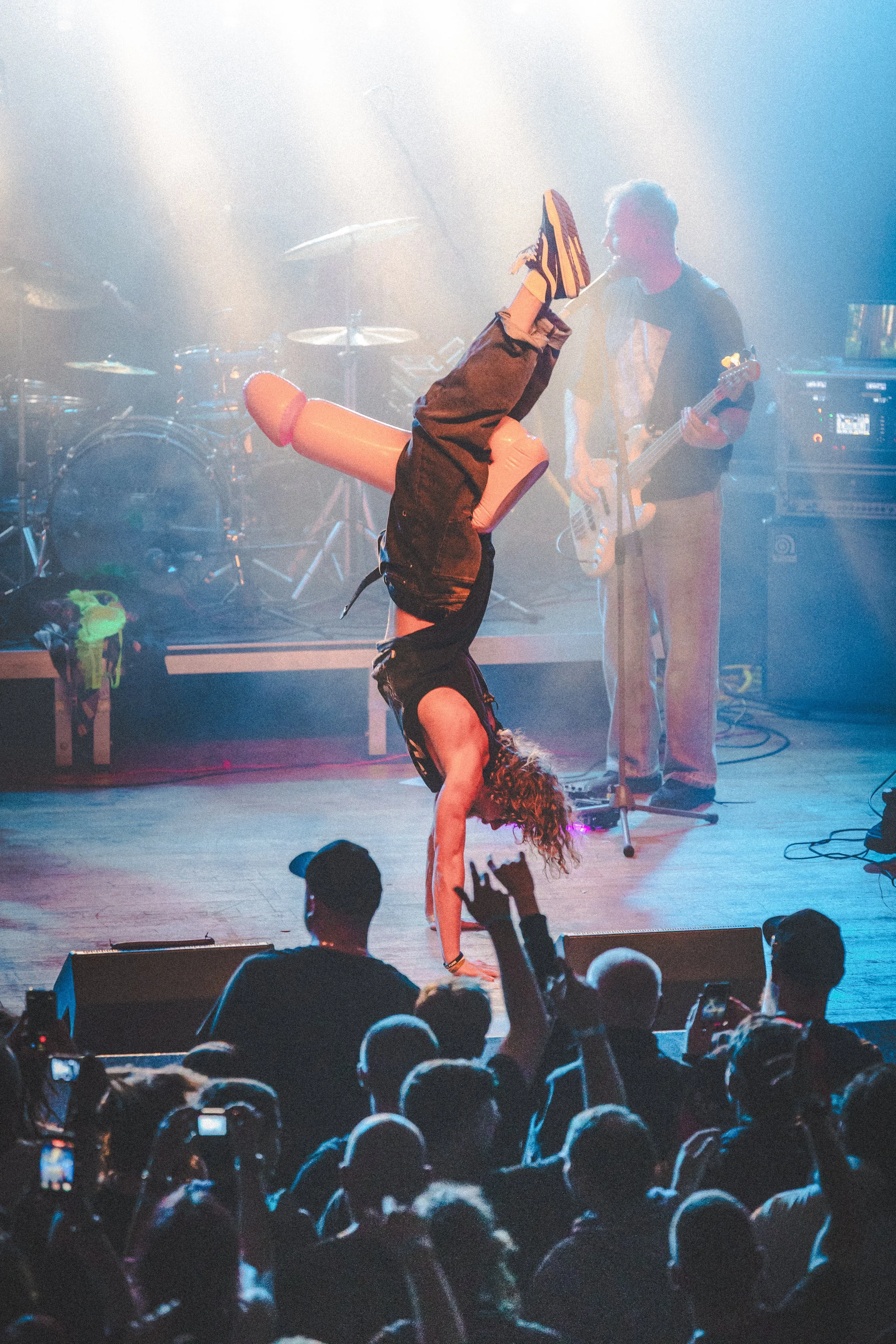 A performer in a handstand with curly hair on stage at a concert, with audience members capturing the moment on phones, and a band playing in the background.
