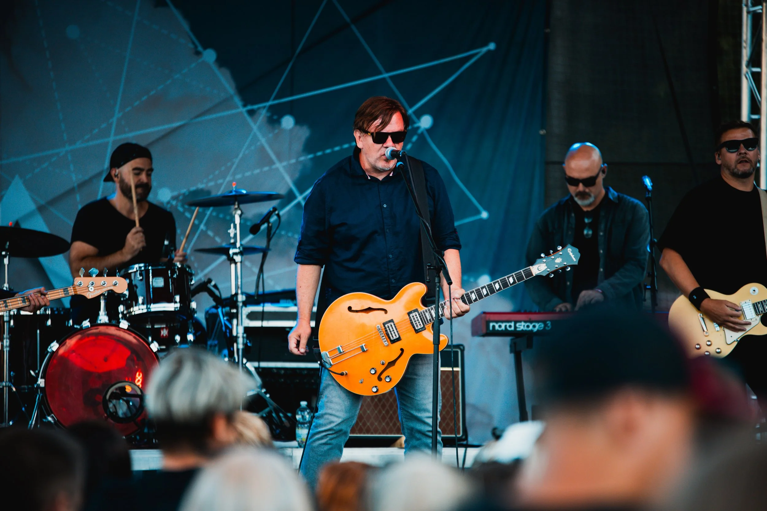 A band performs live on stage, with a man in sunglasses playing an orange guitar at the microphone, drums, keyboard, and other musicians in the background.
