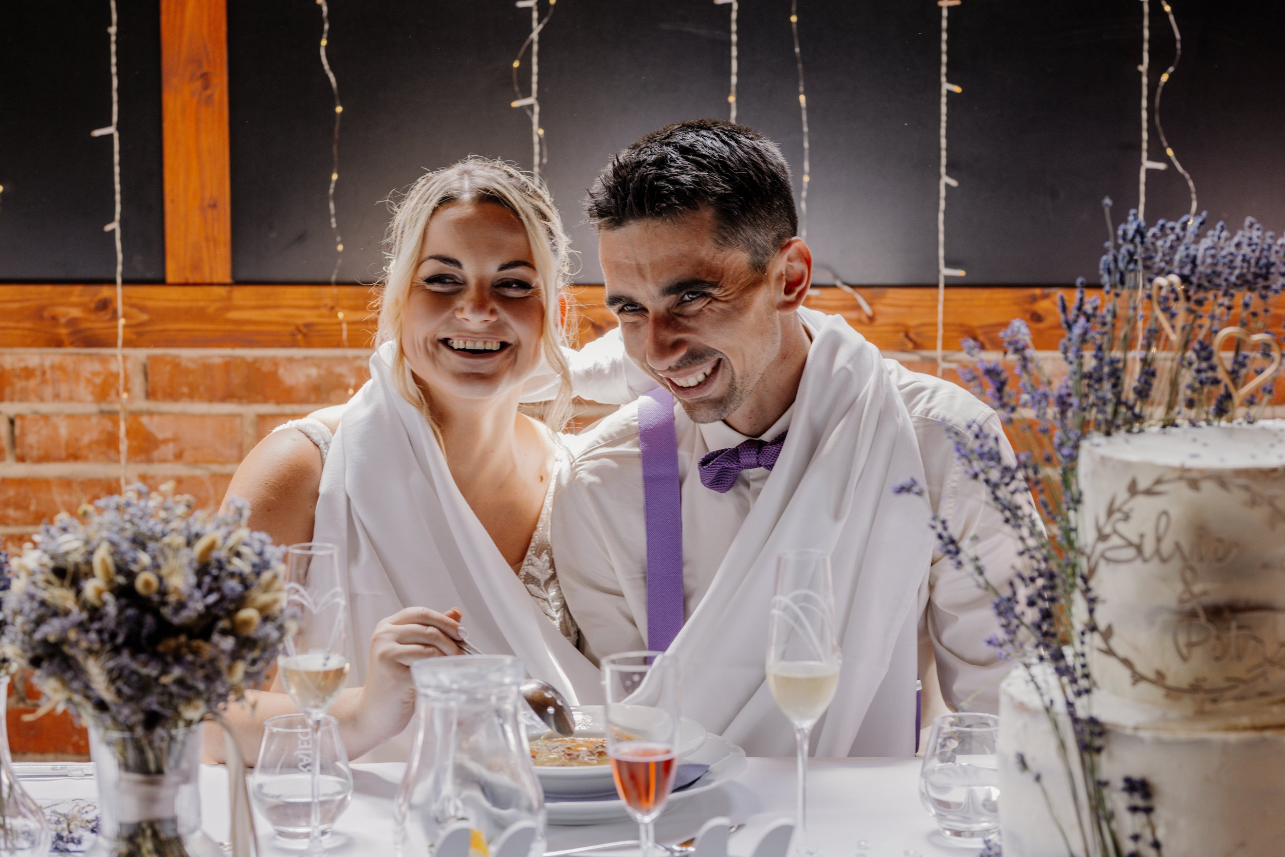 A smiling woman and man sitting at a table during a celebration, with flowers and drinks on the table, and fairy lights in the background.