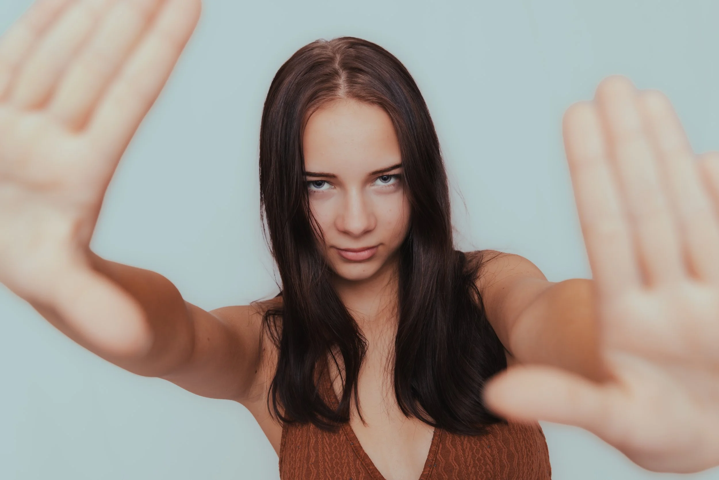 Young woman with long dark hair looking at camera, hands reaching forward, against a light blue background.