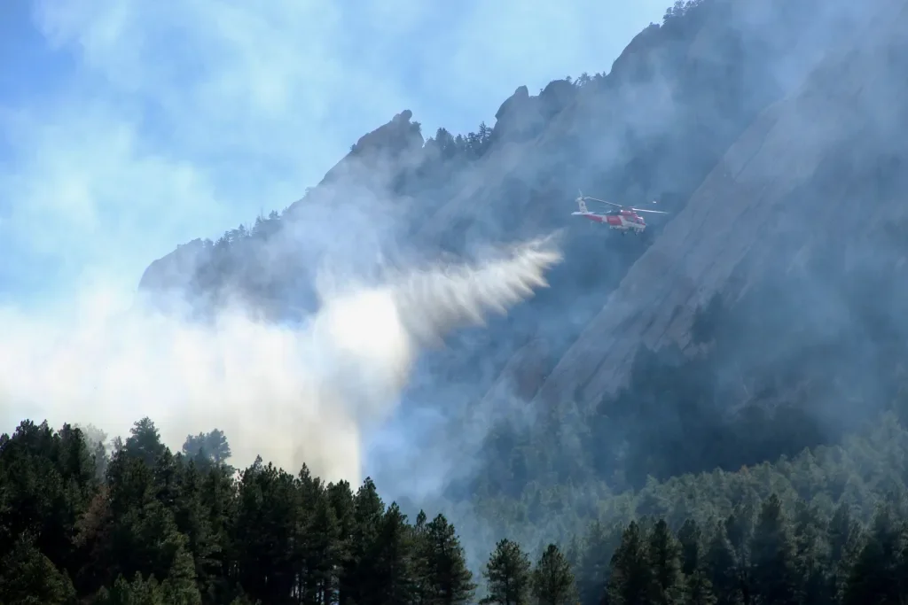 A helicopter flying near a steep mountain, releasing water to combat a forest fire, with smoke rising among trees.