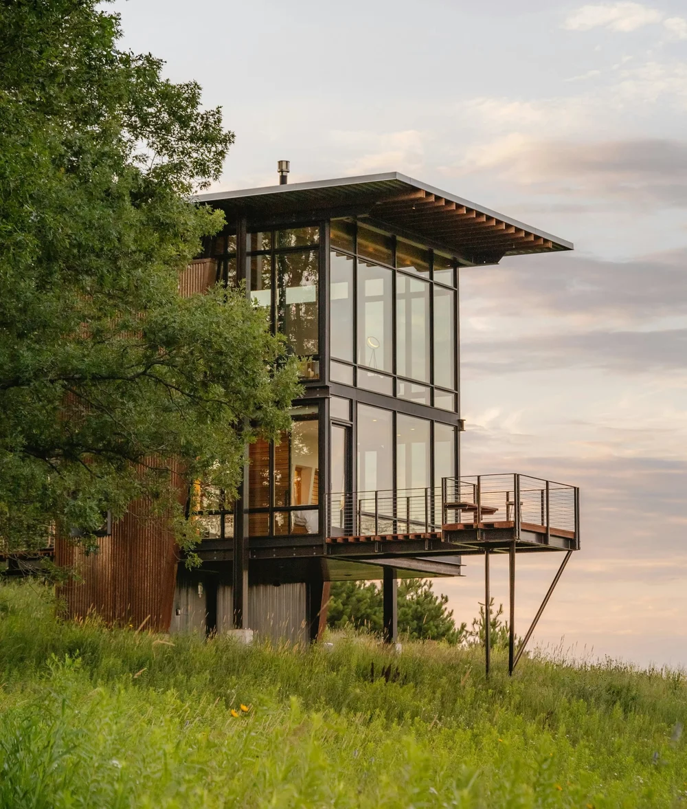 Modern two-story house with large glass windows on a grassy hillside surrounded by trees.