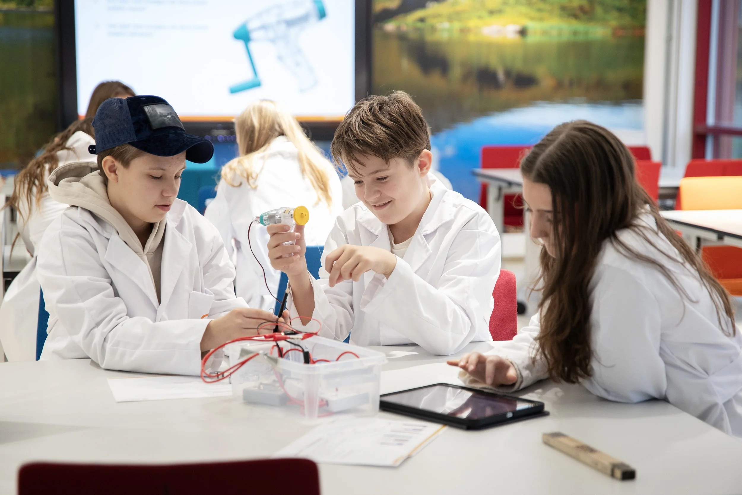 Gruppe ungdommer i hvite labfrakker som jobber med elektriske komponenter i et klasserom.