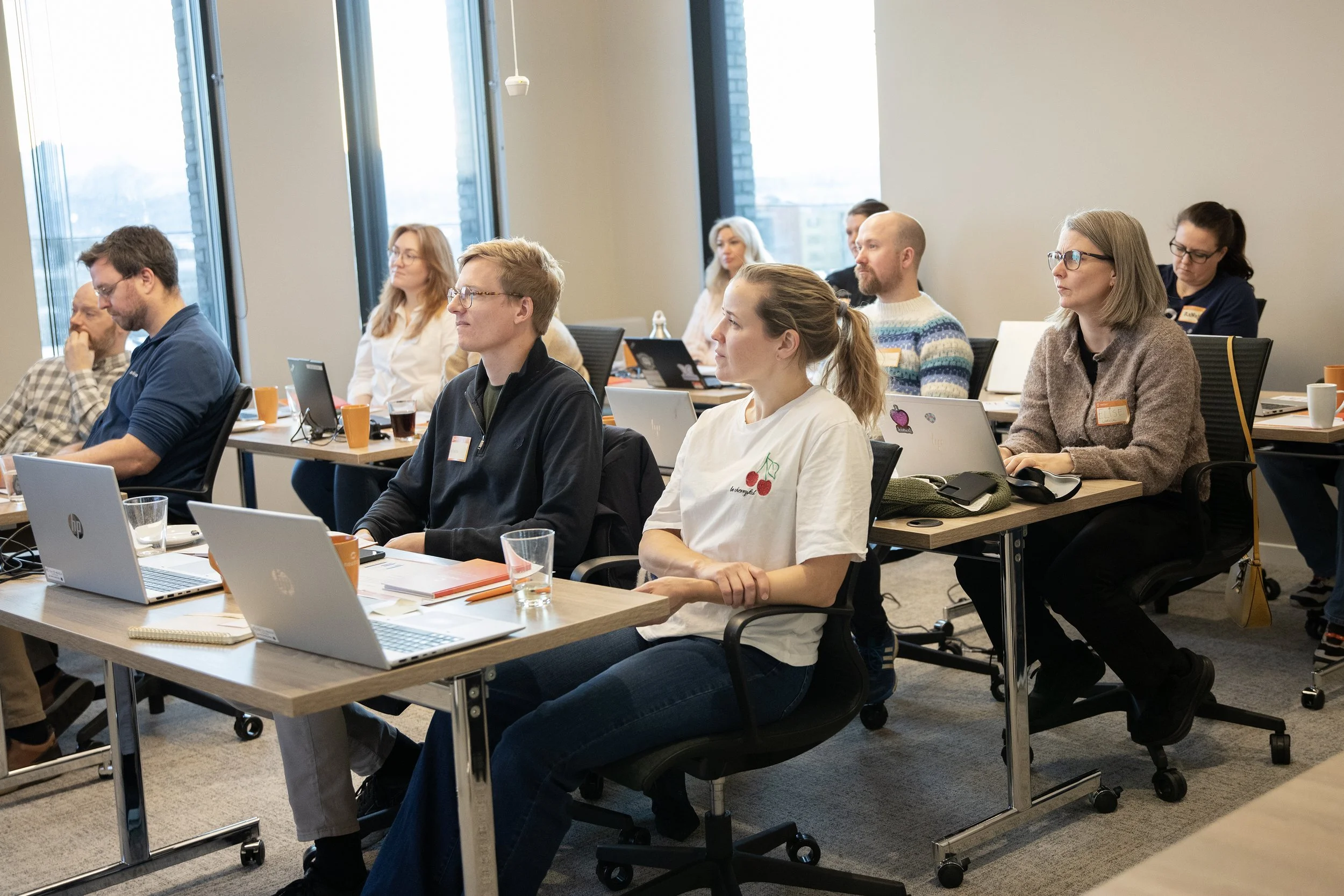 En gruppe mennesker deltar i et møte eller en forelesning i et konferanserom. De sitter ved bord med åpne laptops, papirer og drikker. Noen ser ut til å være konsentrerte og lytte, mens andre jobber på sine enheter.