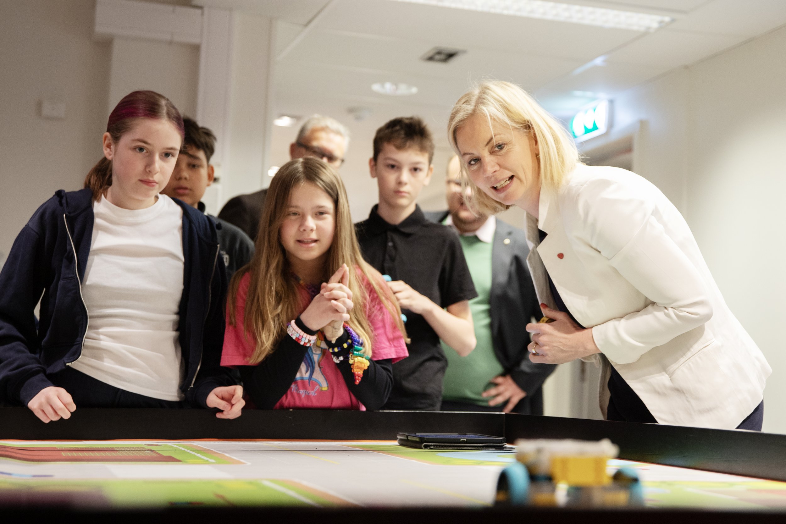 En gruppe mennesker, inkludert unge jenter og voksne, står rundt et bord med et utendørs spill eller aktivitet. En kvinne i hvit frakk snakker med gruppen. De ser fokusert og engasjerte ut.