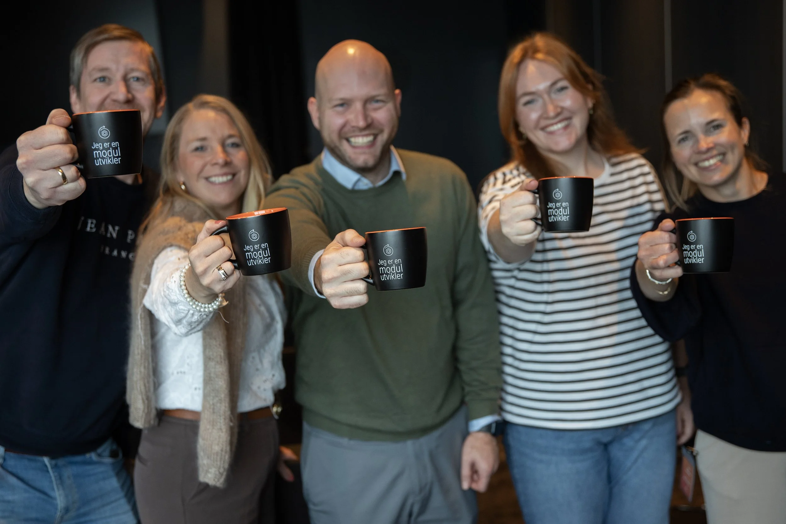 Fem personer smiler og holder opp svarte krus med teksten 'Jeg er en modul utvikler' i en gruppe, inne i et rom.