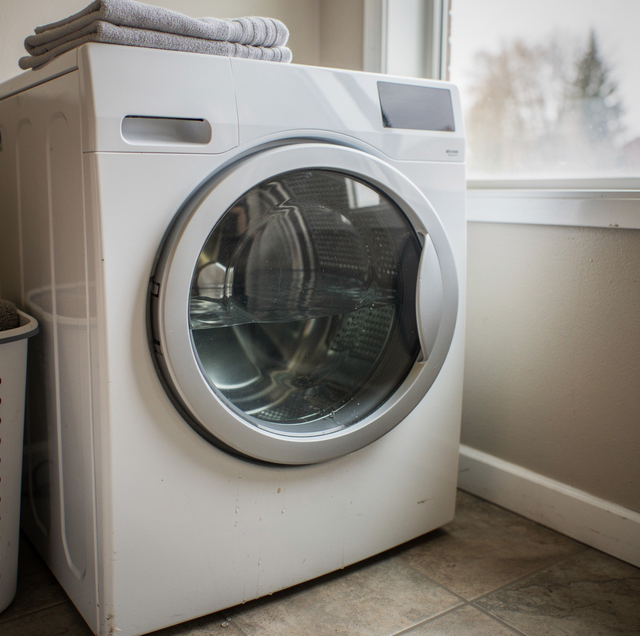A white front-load washing machine with folded towels stacked on top sits next to a window in a laundry room.