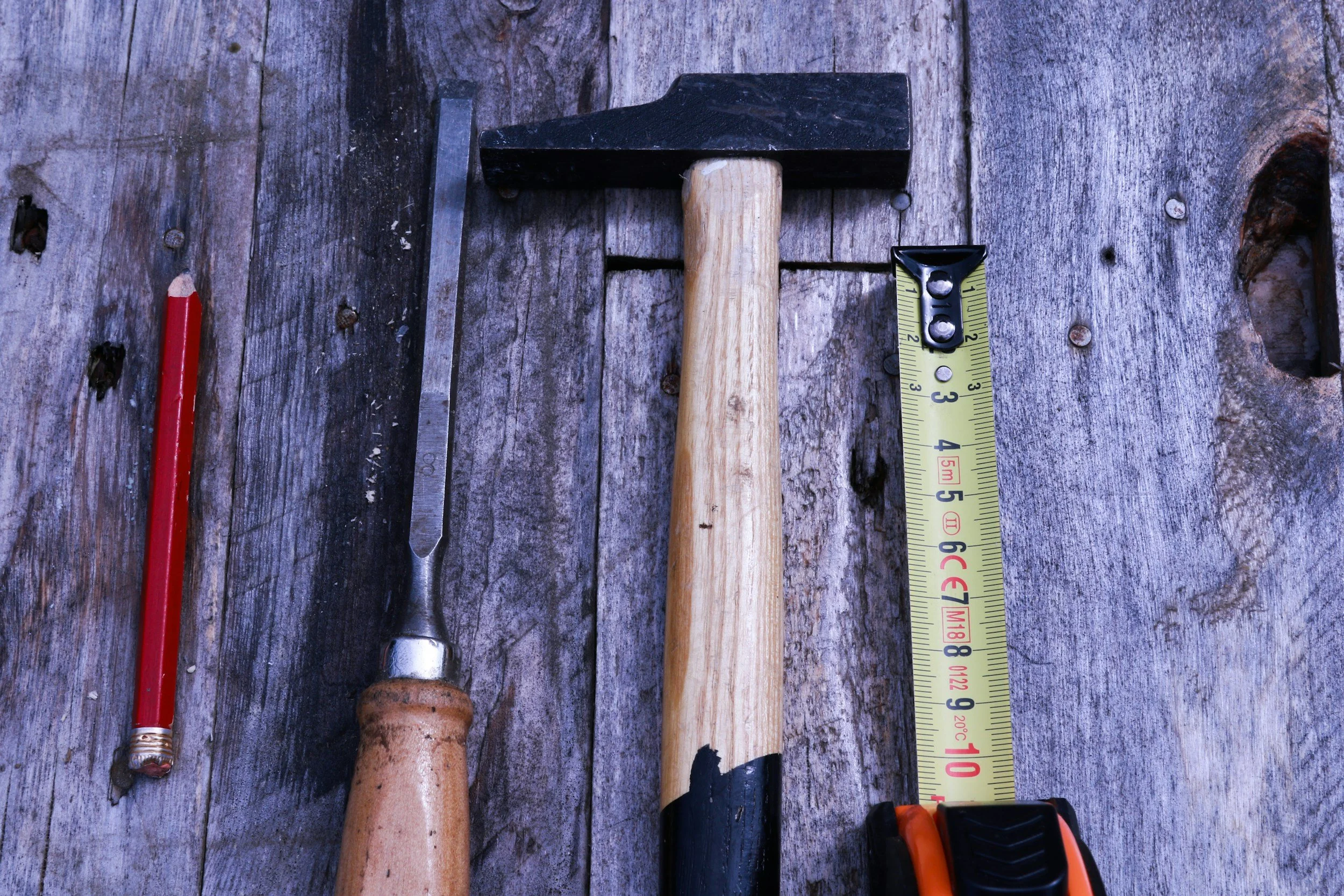 A variety of hand tools on a weathered wooden surface, including a red pencil, a metal file, a small hammer with a wooden handle, a tape measure, and a power drill.