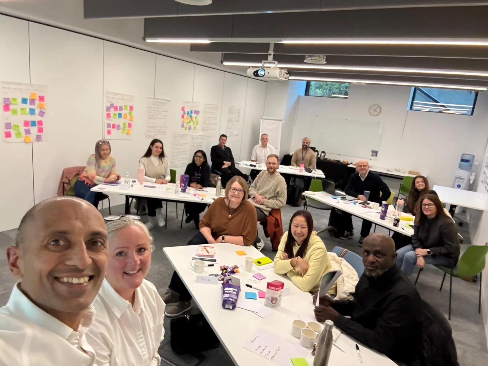 A group of diverse people in a classroom or conference room, seated at tables with notebooks, coffee cups, snacks, and writing materials, smiling for a group selfie with posters and papers on the wall behind them.
