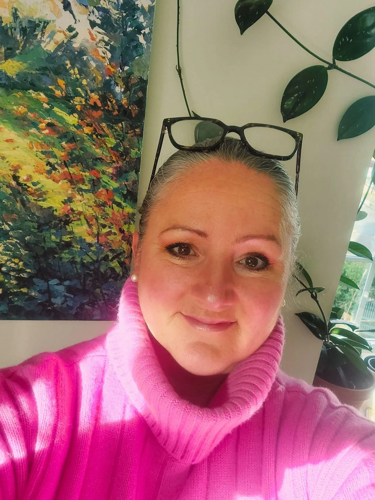 A woman with silver hair wearing a pink turtleneck sweater, smiling, with glasses resting on her head. In the background, there's a colorful painting aside green houseplants and a window.