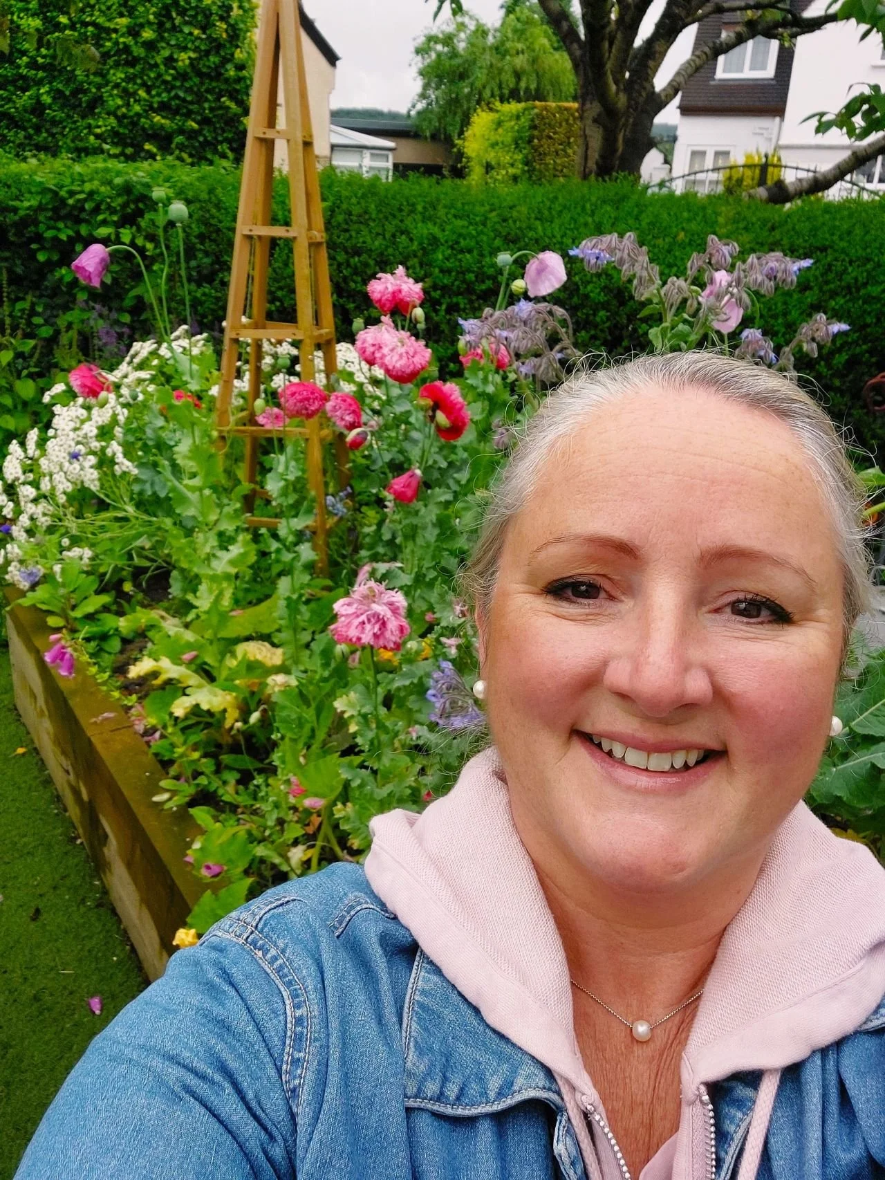 A smiling woman taking a selfie in a garden with colorful flowers, green bushes, trees, and houses in the background.