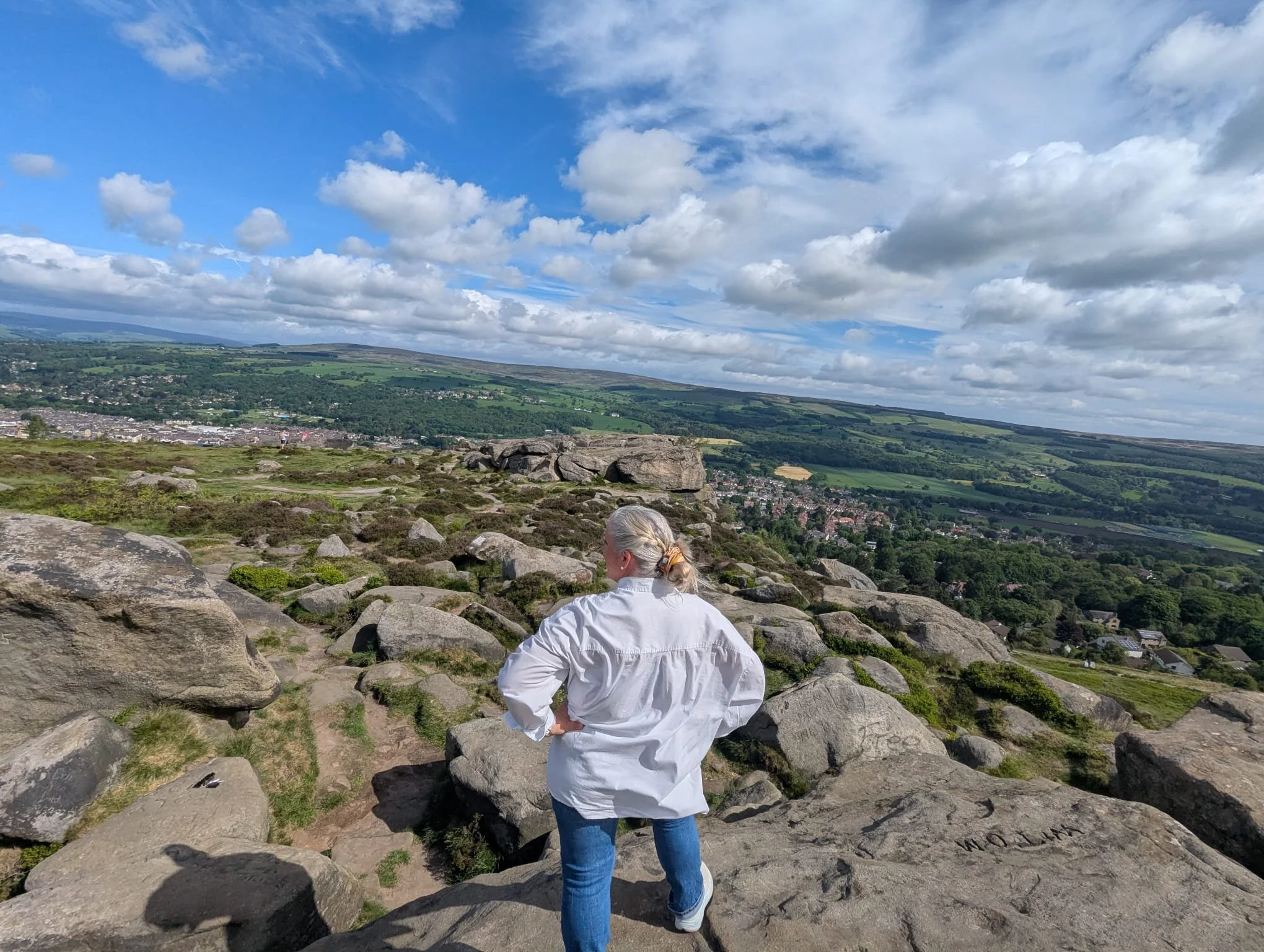 A woman with gray hair tied back, wearing a white jacket and jeans, stands on rocky terrain overlooking a green valley with houses, fields, and hills under a partly cloudy sky.