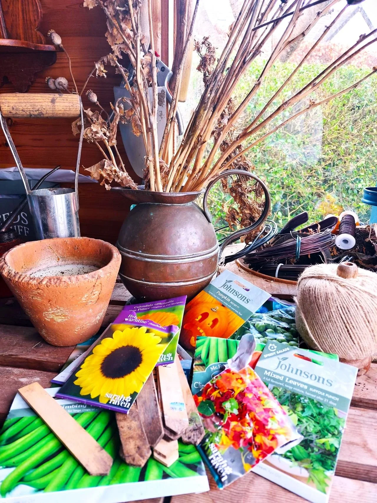 A cluttered rustic wooden table with gardening and planting supplies, seed packets, dried stalks in a metal pitcher, a terracotta pot, gardening tools, a ball of twine, and a window with greenery outside.
