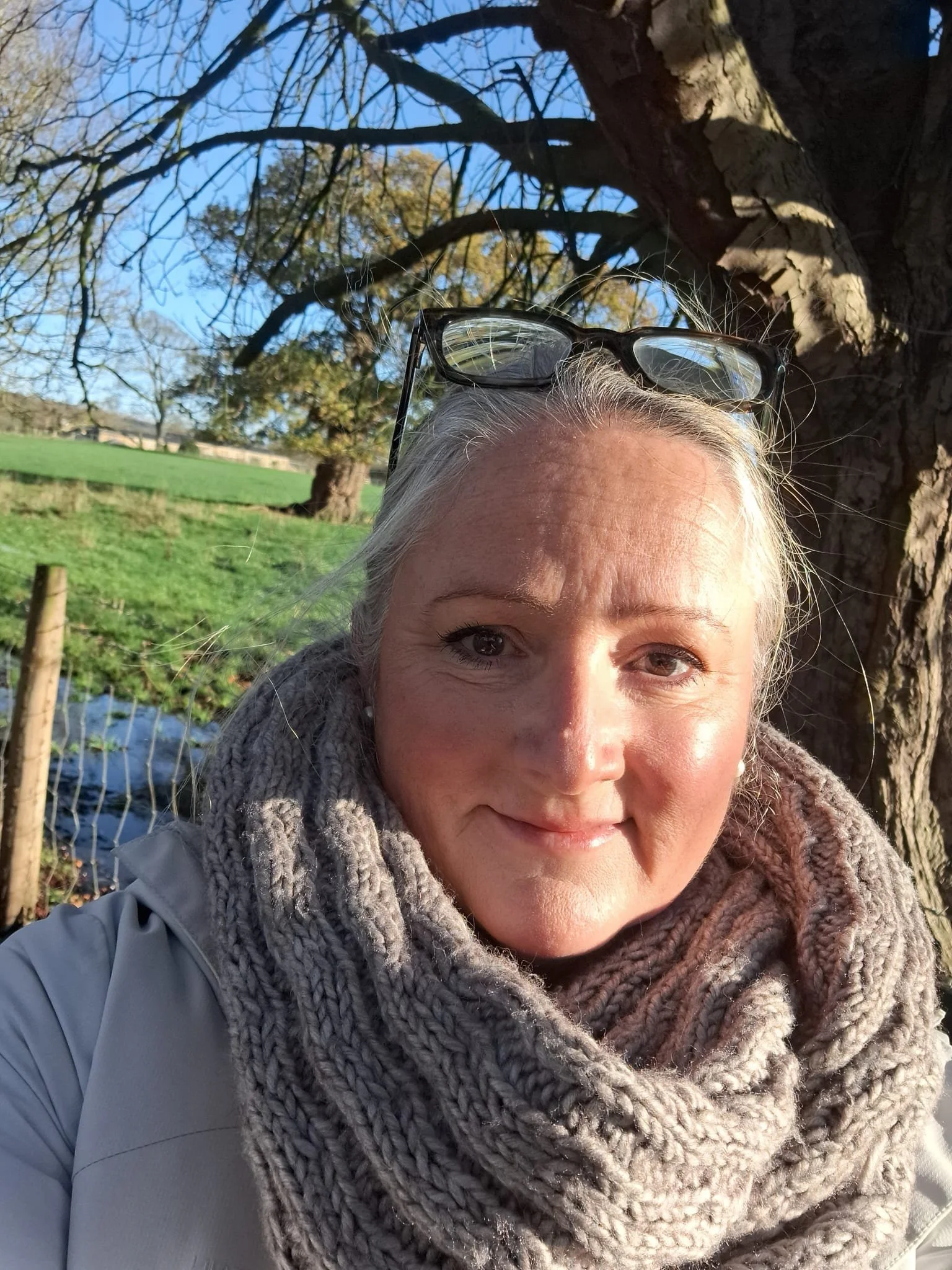 Close-up of a woman with glasses resting on her head, wearing a thick knitted scarf, outdoors with trees and a field in the background on a sunny day.