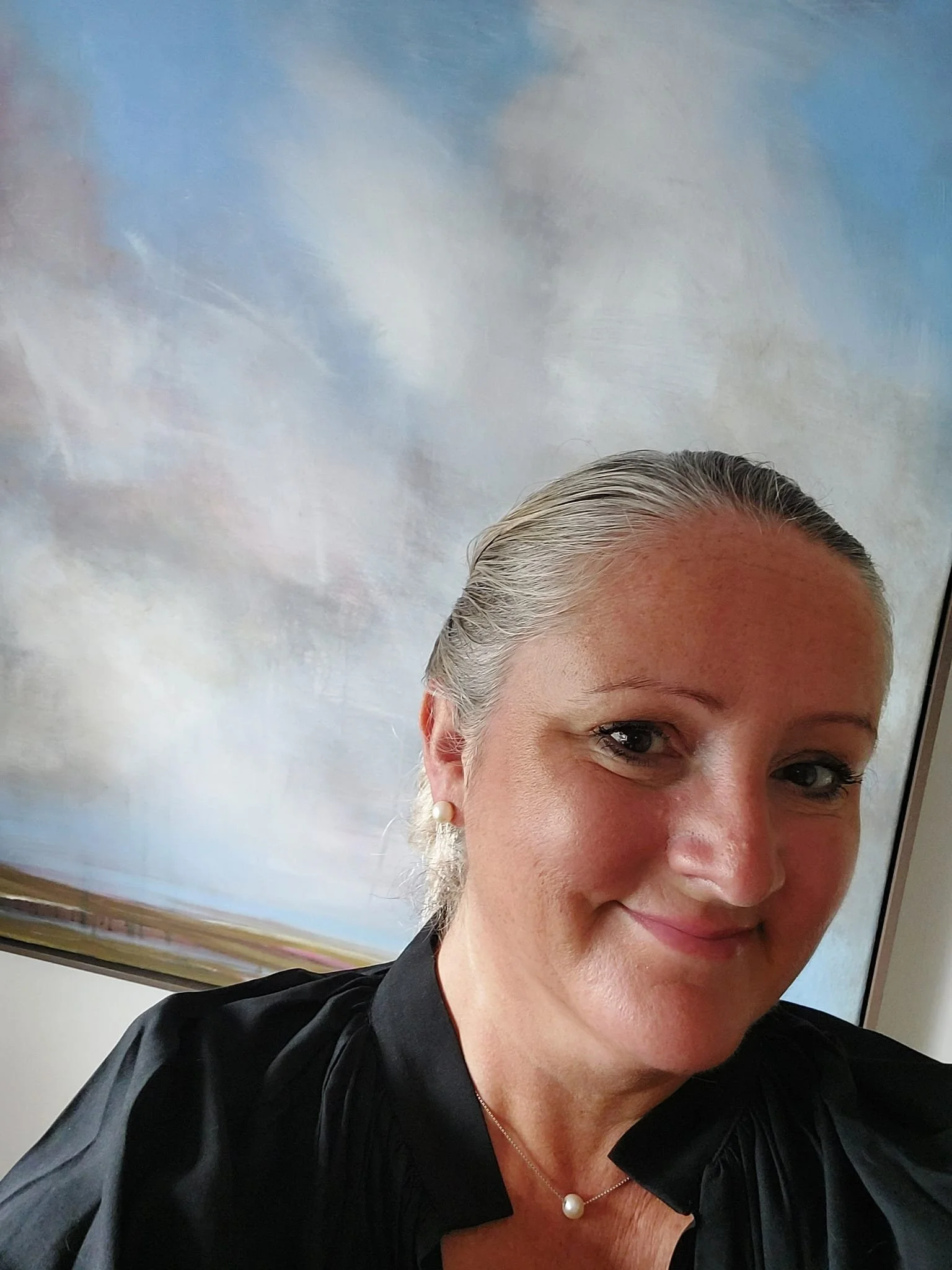 A woman with gray hair tied back, smiling, wearing earrings and a pearl necklace, inside an airplane near a window showing cloudy sky.