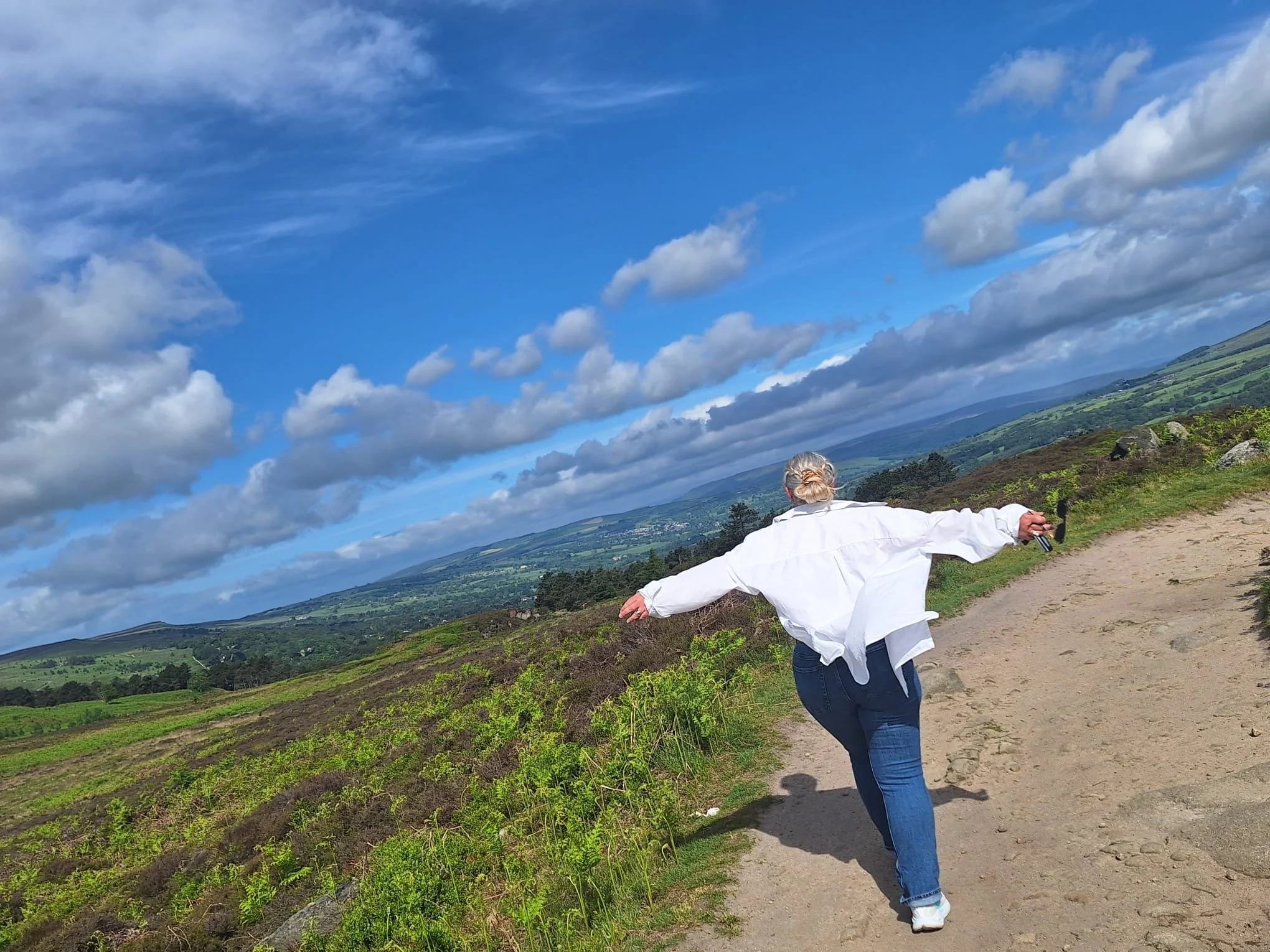 A woman with gray hair is enjoying a scenic view while standing outdoors on a trail with her arms outstretched. The landscape features green rolling hills, open fields, and a partly cloudy sky.